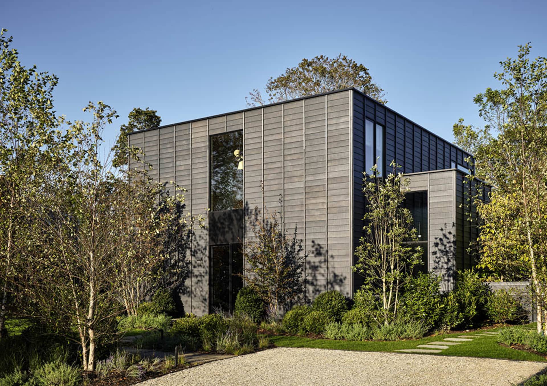 A modern, metal-clad house with large windows and a gravel driveway surrounded by greenery and young trees.