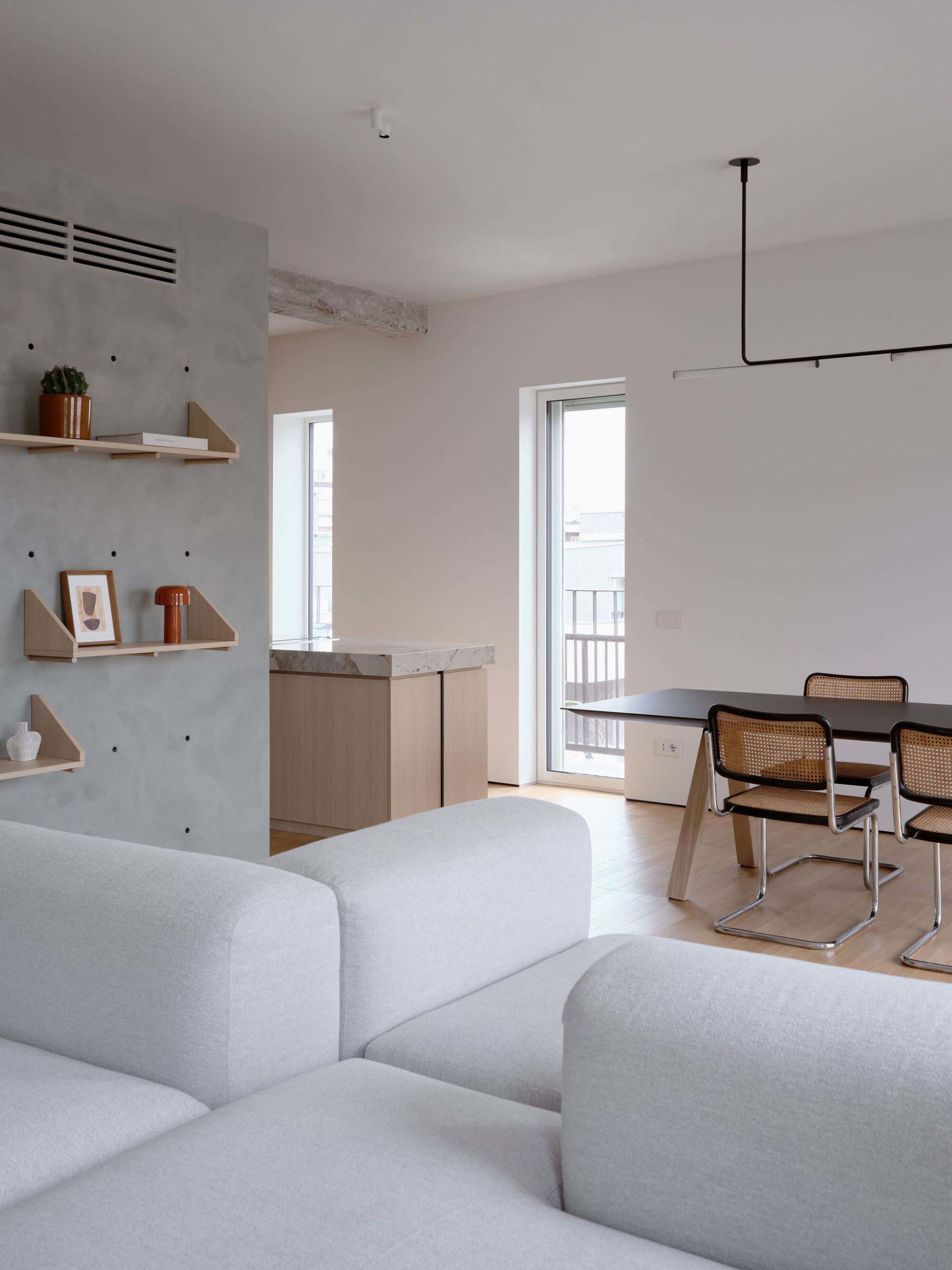 A minimalist living room with concrete walls, a white sofa, and a dining area with a black table and bamboo chairs.