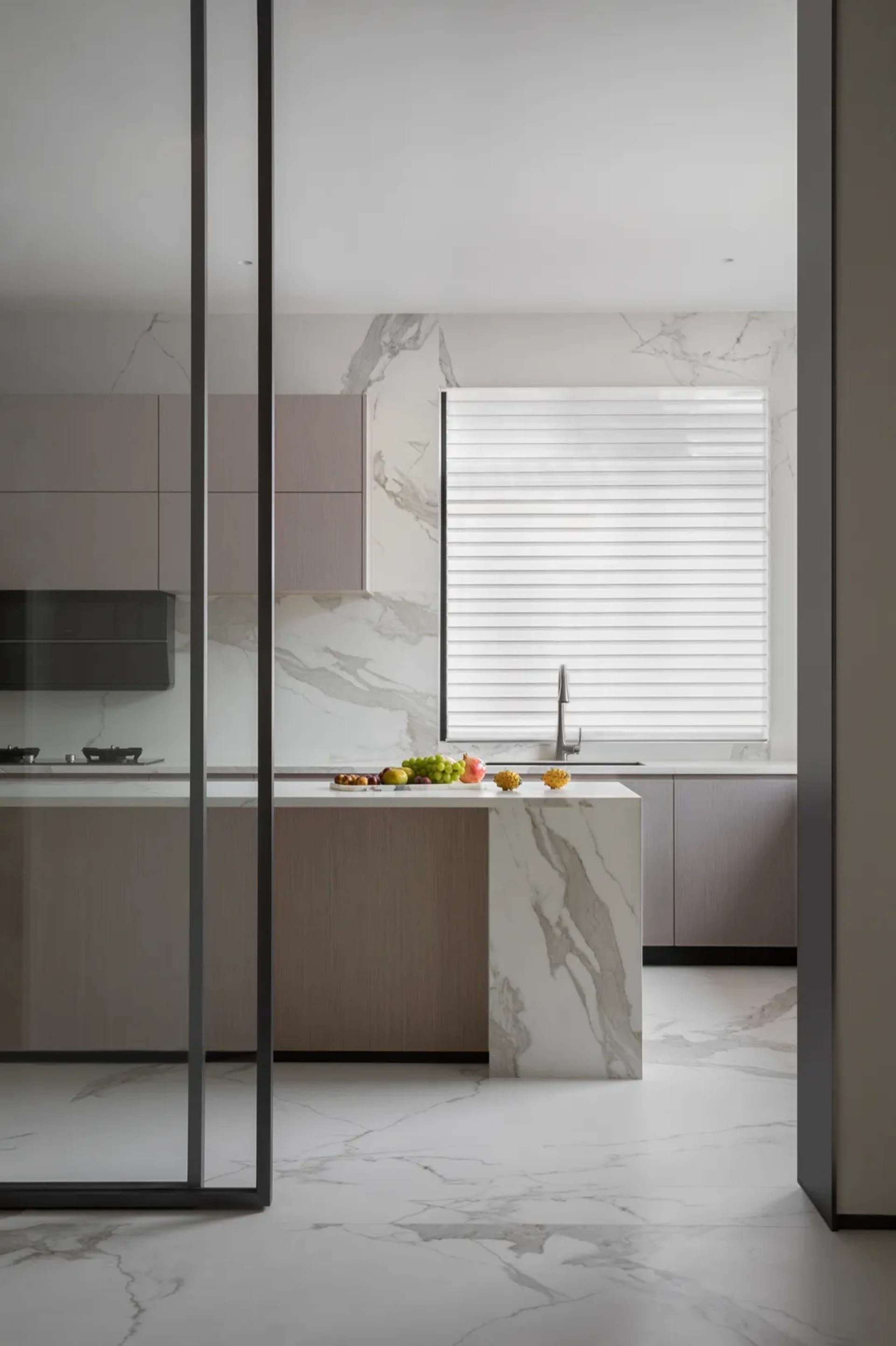 A minimalist residential kitchen with marble countertops and floor, featuring a kitchen island and a window with blinds.