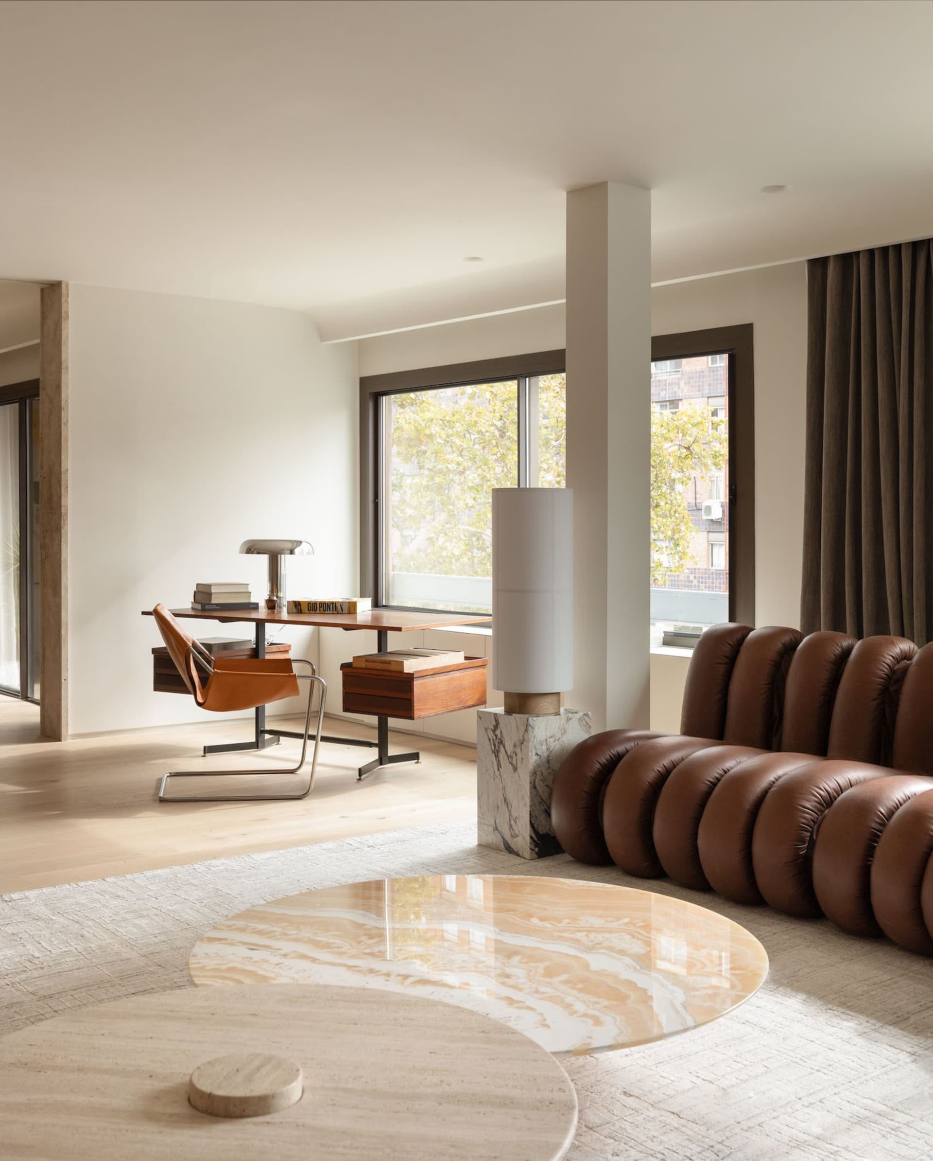 A minimalist living room with a round marble coffee table, brown leather sofa, and wooden desk.