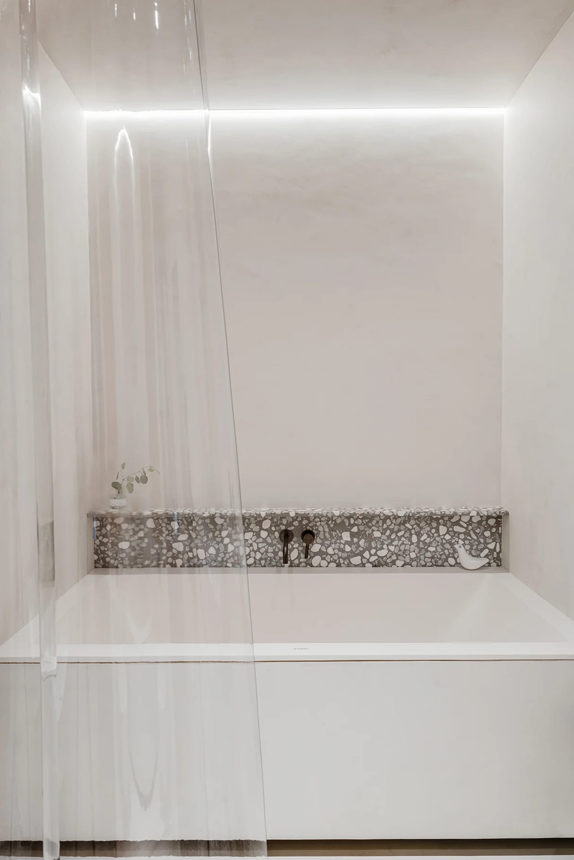 Modern minimalist bathroom featuring a white bathtub with a textured terrazzo backsplash and illuminated lighting.