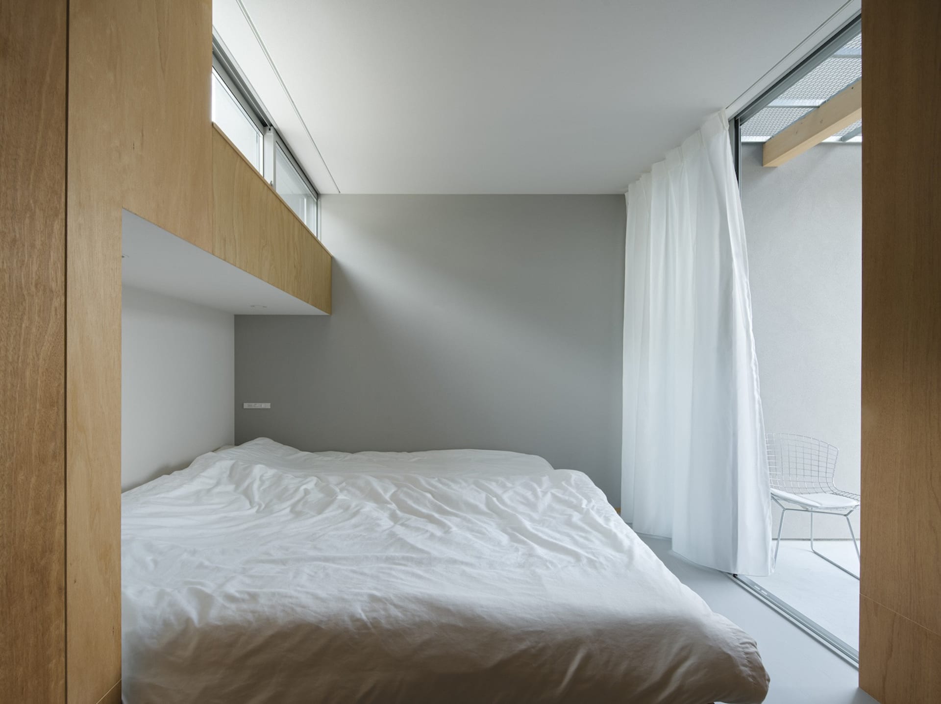 A minimalist bedroom with a white bed, white curtains, and light wooden walls.