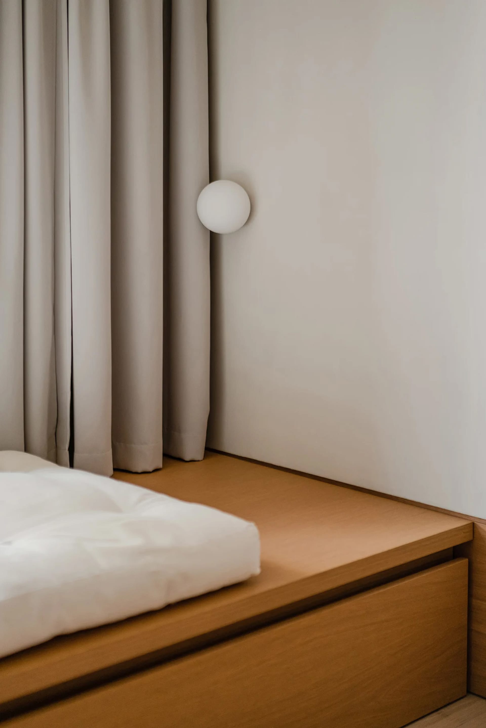 A minimalist bedroom featuring a wood-framed bed against a neutral-colored wall with a simple, spherical light fixture.