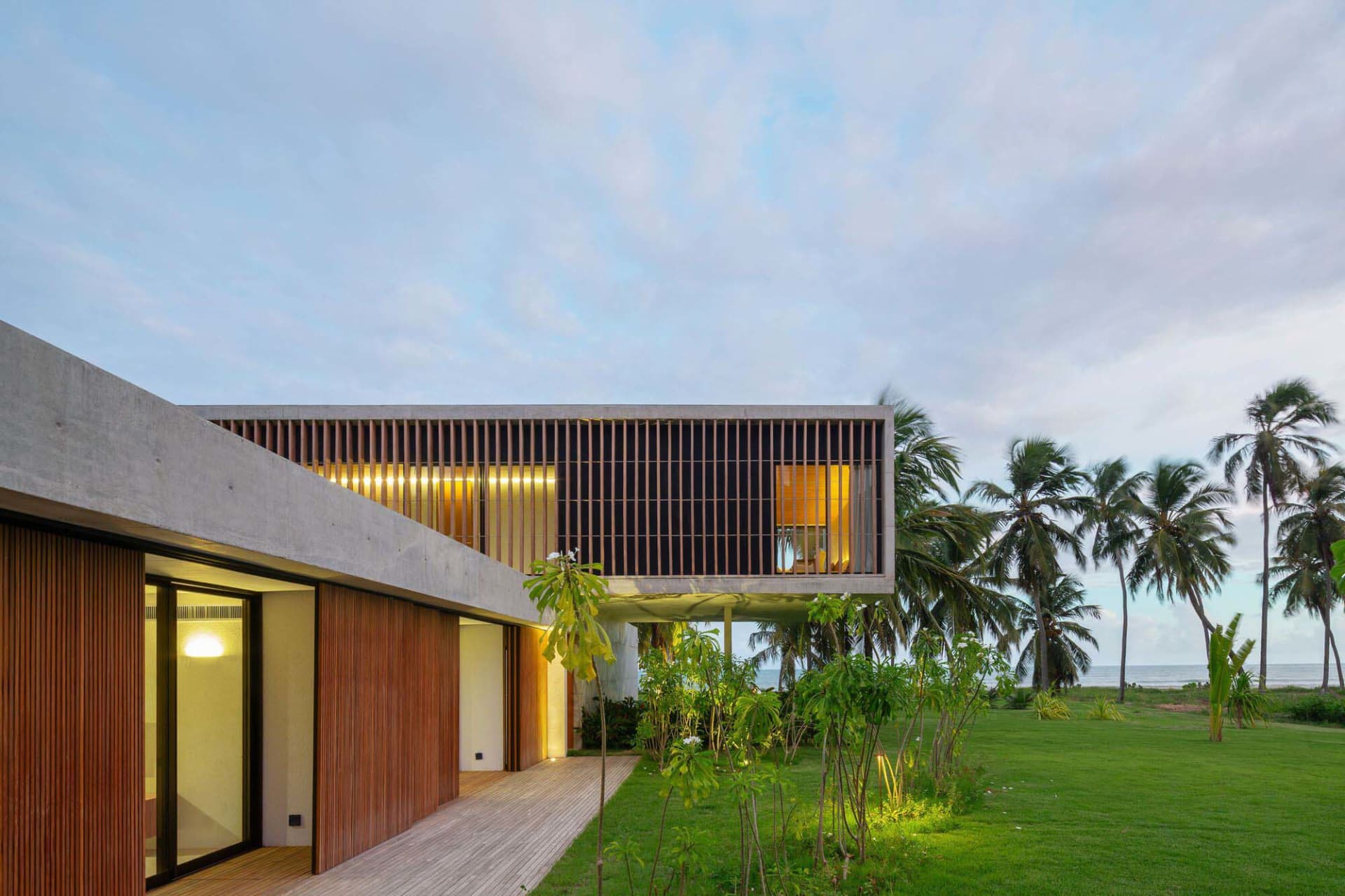 A modern house featuring concrete, glass, and wooden elements with a spacious lawn and palm trees.