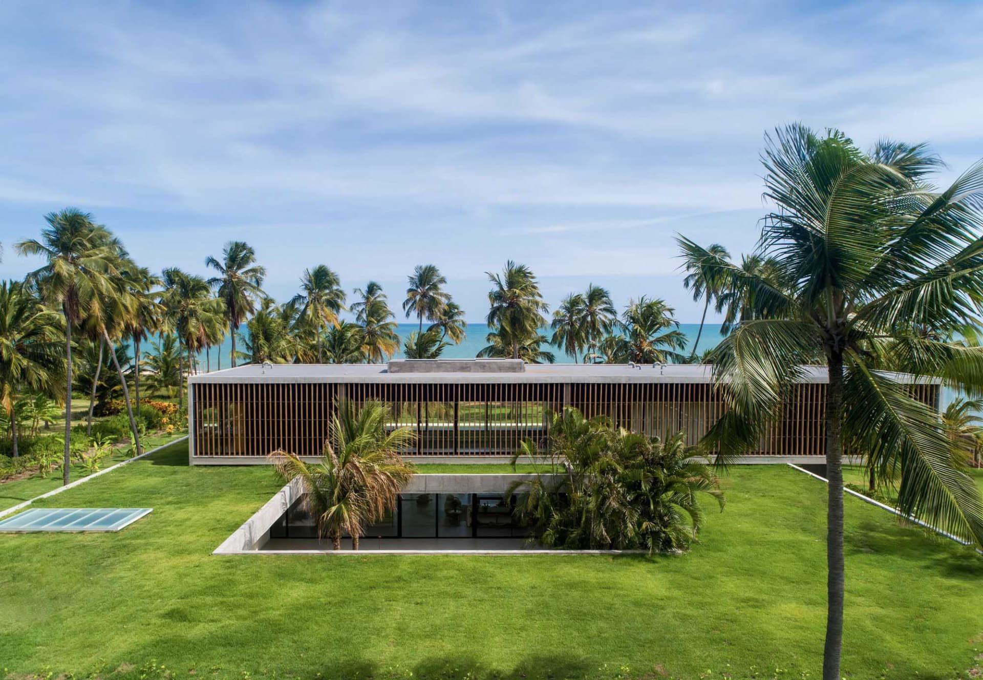 A modern house with a concrete exterior surrounded by lush greenery and palm trees, overlooking the ocean.