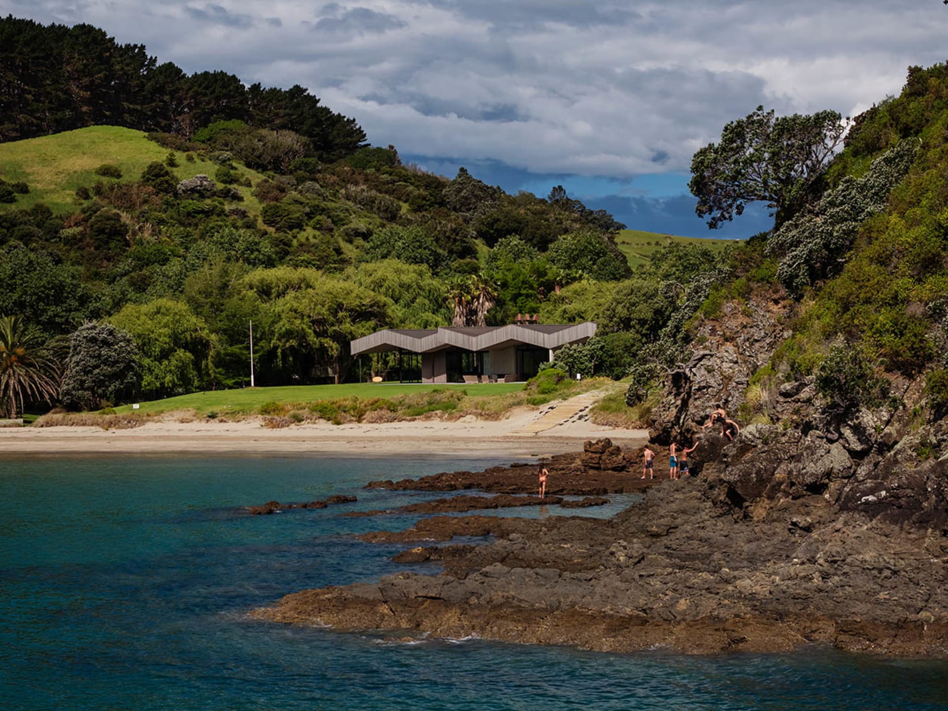 Modern beach house with concrete, wood, and tiled flooring, surrounded by lush greenery.