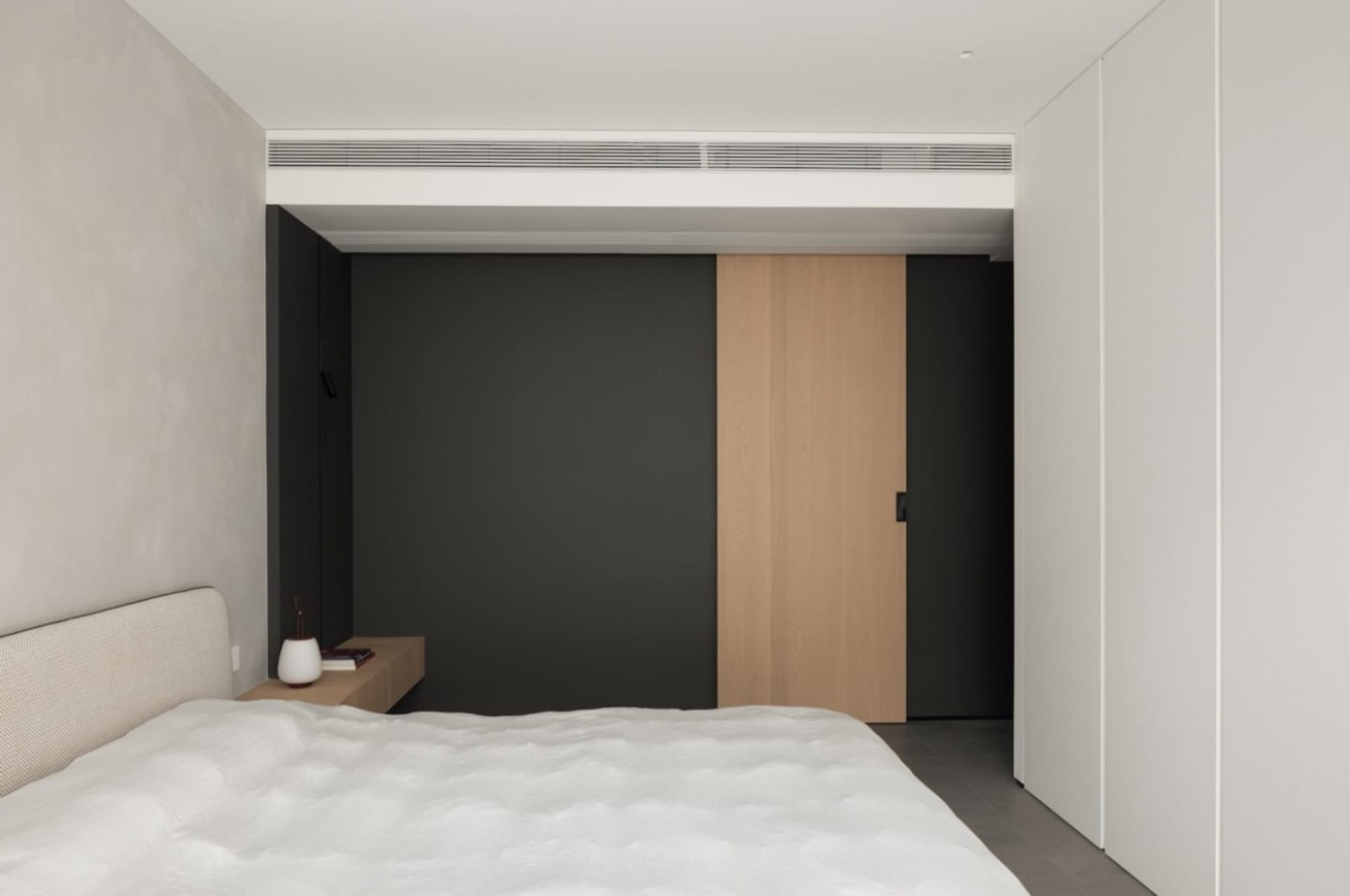 A modern minimalist bedroom featuring a white bed with a tufted headboard, a small wooden nightstand, and a sliding door with