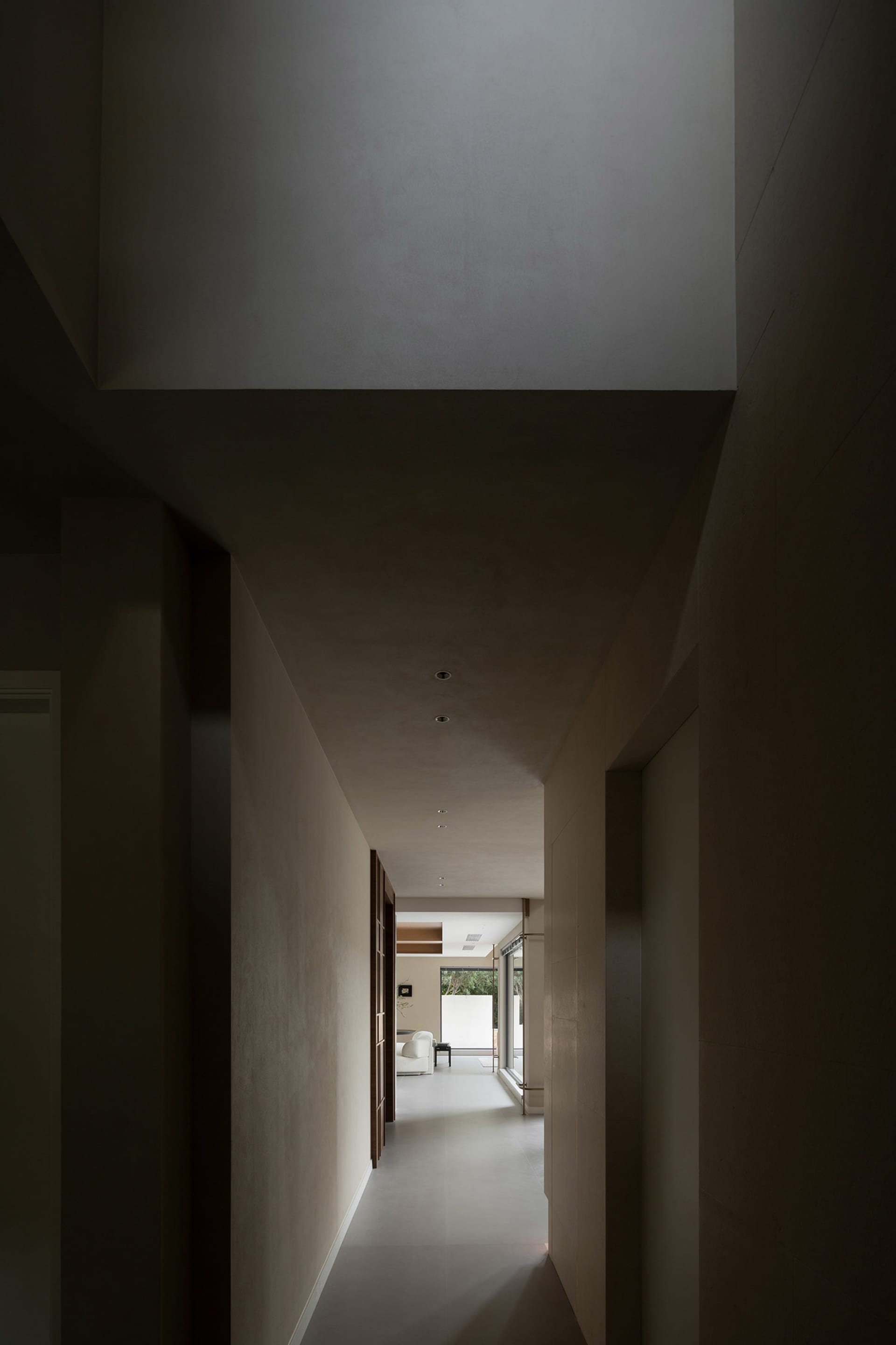 A minimalist interior corridor with concrete walls and white flooring, leading to a brightly lit room with a white couch and