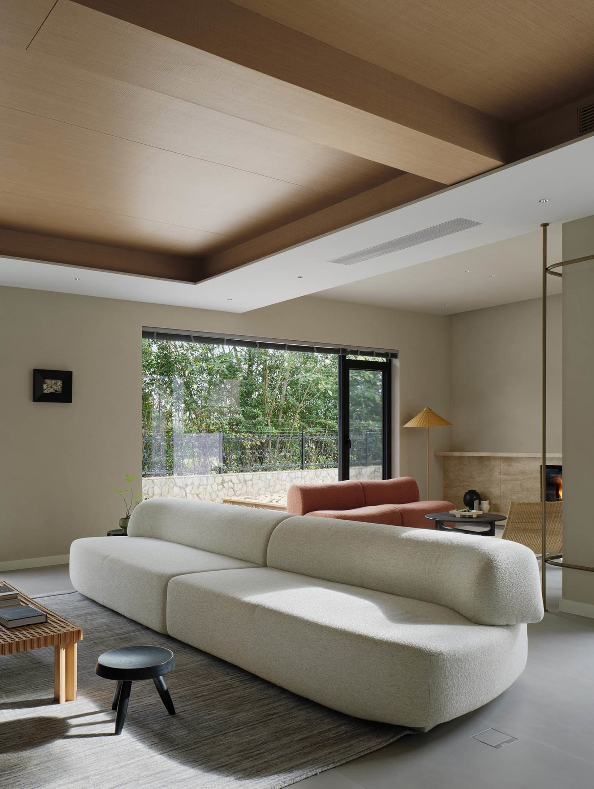 A modern residential interior living room featuring a white sofa, a wooden coffee table, a black stool, and a large window ov