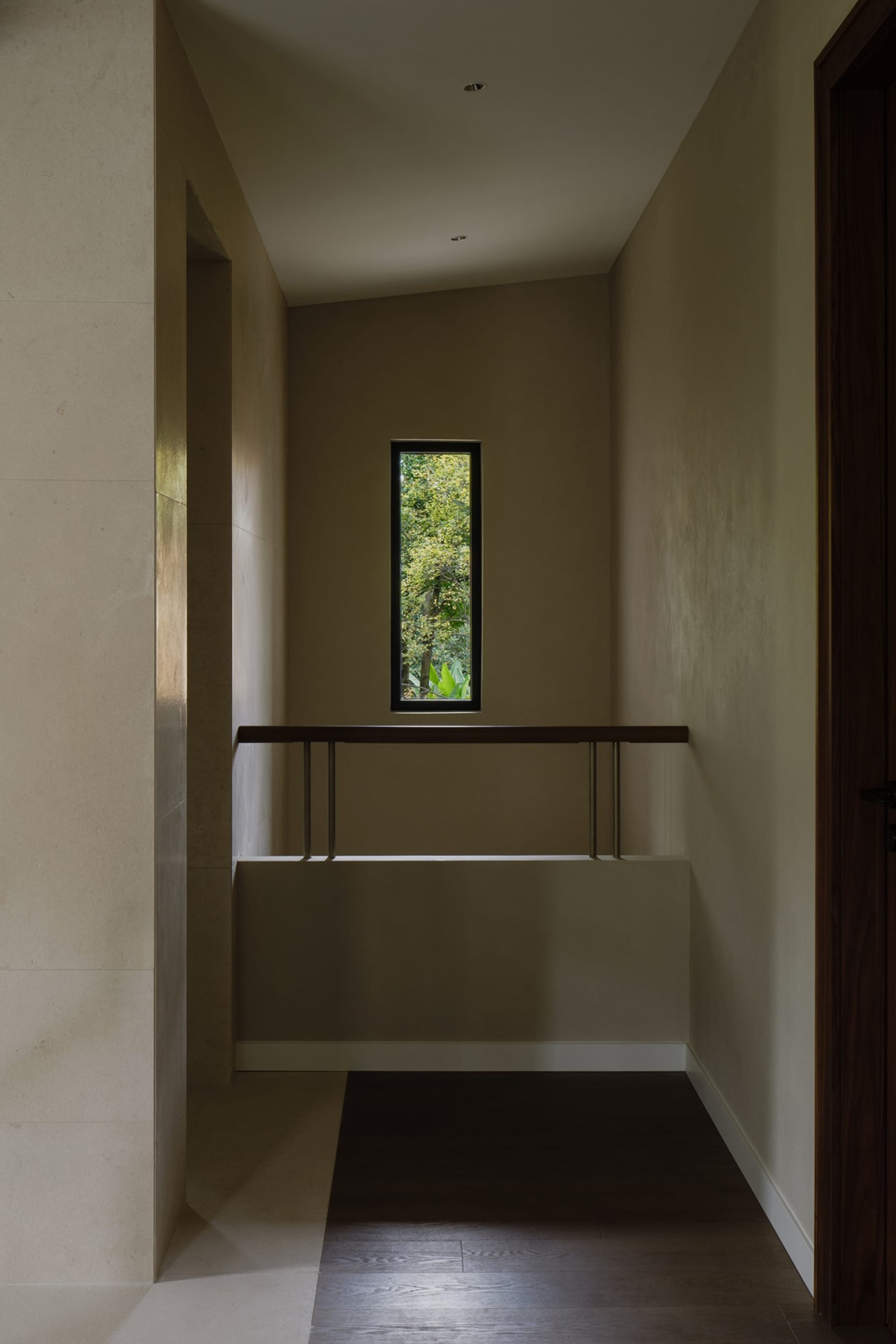 A corridor with neutral-colored walls and floors, featuring a window with a view of greenery and a wooden railing.