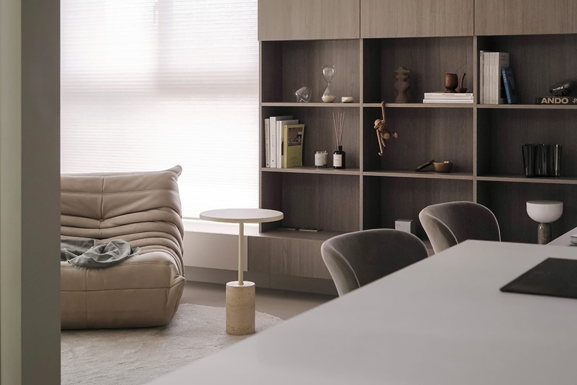 A minimalist living room featuring a beige leather sofa, a white round side table, and a wooden shelving unit with books and 