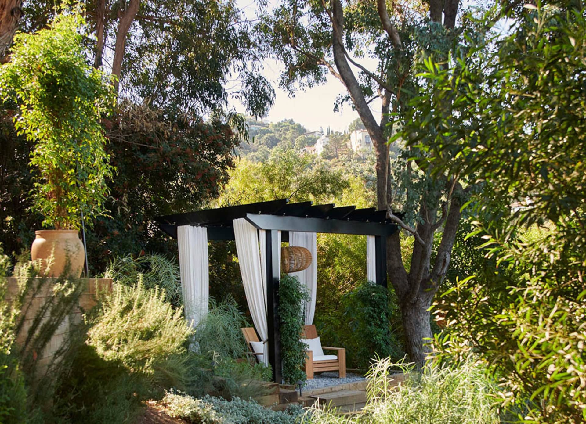 A modern pavilion with white curtains and a wooden chair surrounded by lush greenery and trees.
