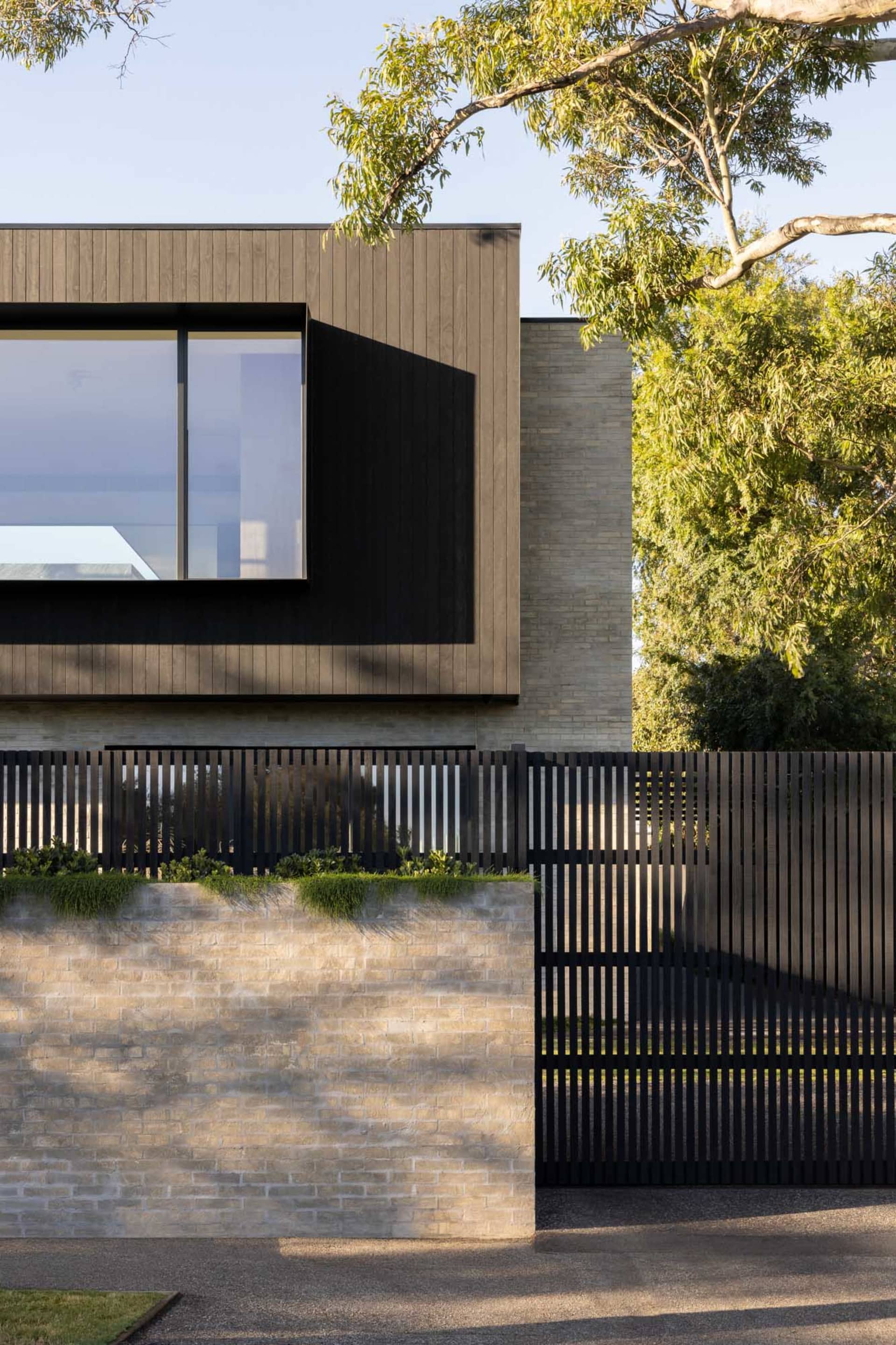 A modern house exterior with brick and wooden paneling, accompanied by a wooden fence and lush greenery.