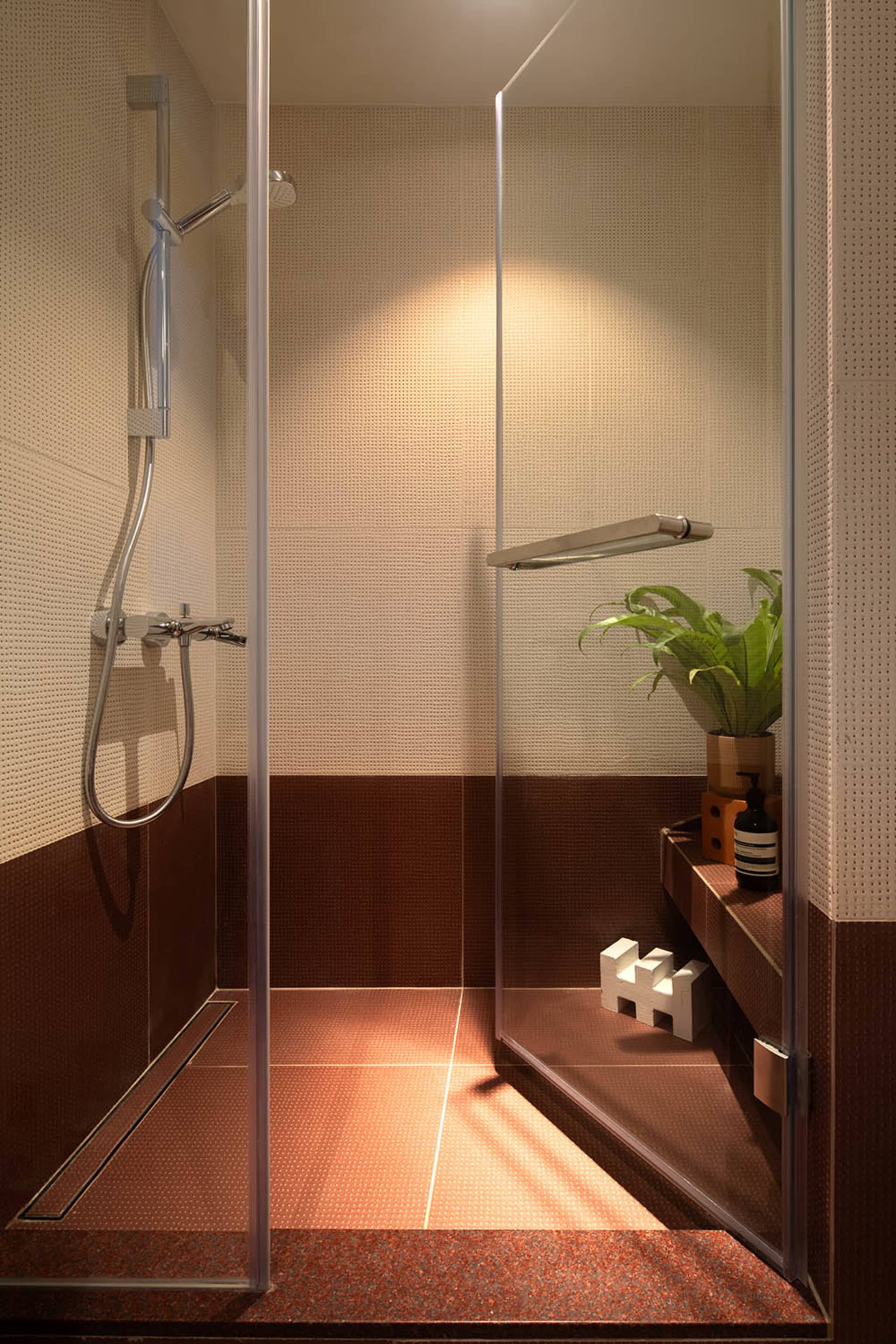 A minimalist bathroom featuring red tiles on the lower half of the walls, with a glass shower enclosure and a potted plant.