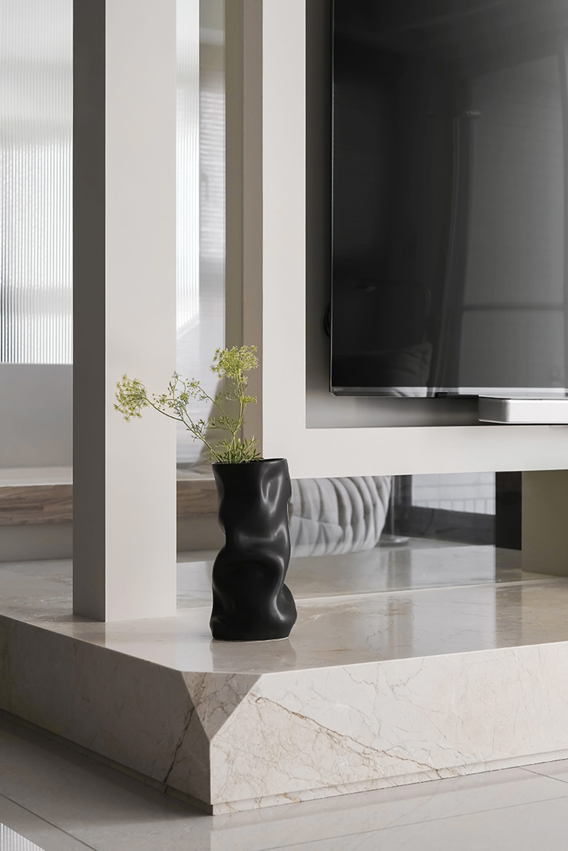A minimalist living room with marble and concrete accents, featuring a black vase with green flowers on a marble countertop.