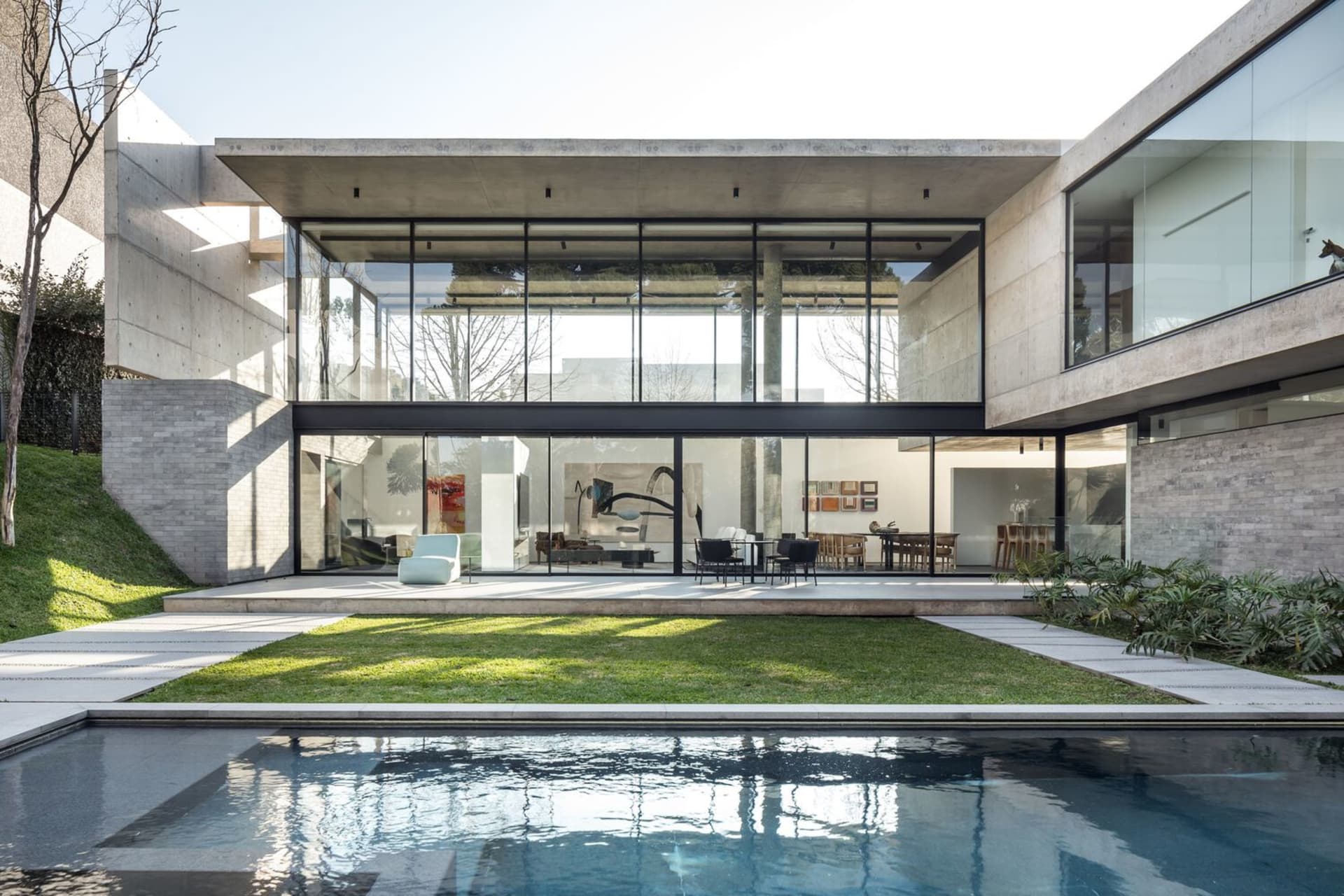 A modern residential home with concrete walls and glass walls and doors, featuring a swimming pool and a large green lawn.