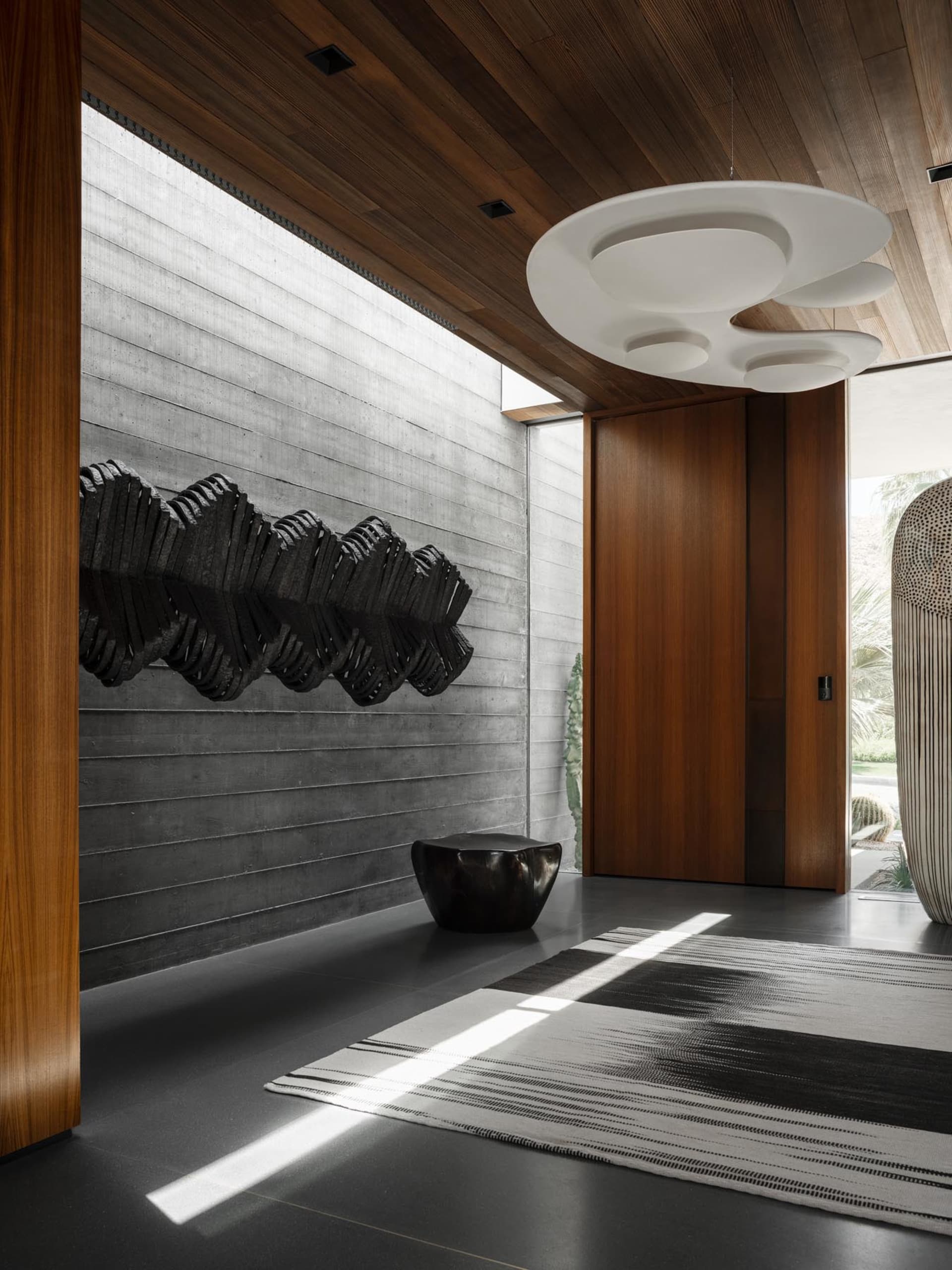 A modern entryway features concrete walls, wooden ceiling beams, and a unique hanging light fixture.