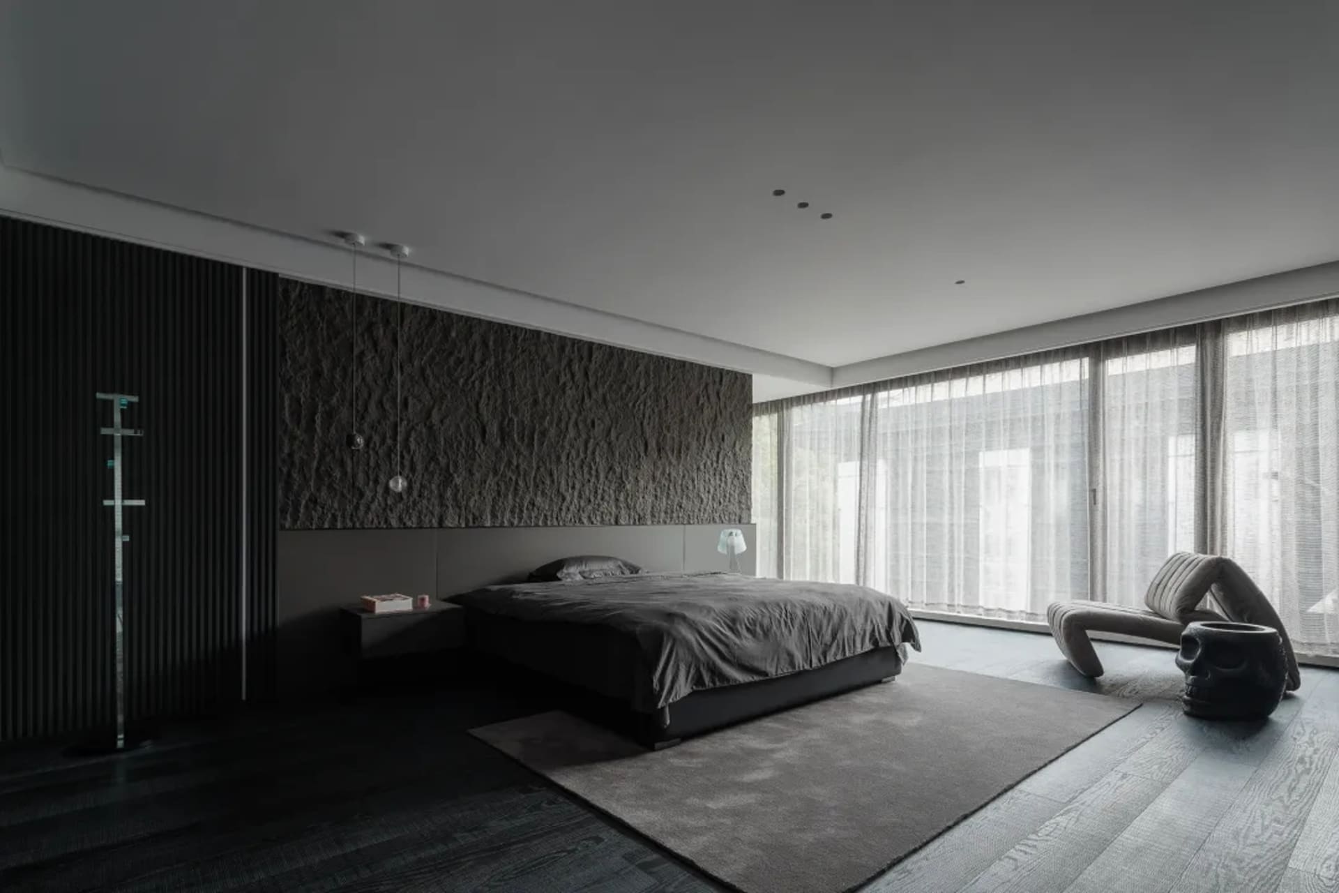 A minimalist bedroom featuring dark wood floors, stone accent wall, and glass walls with a bed and wooden furniture.