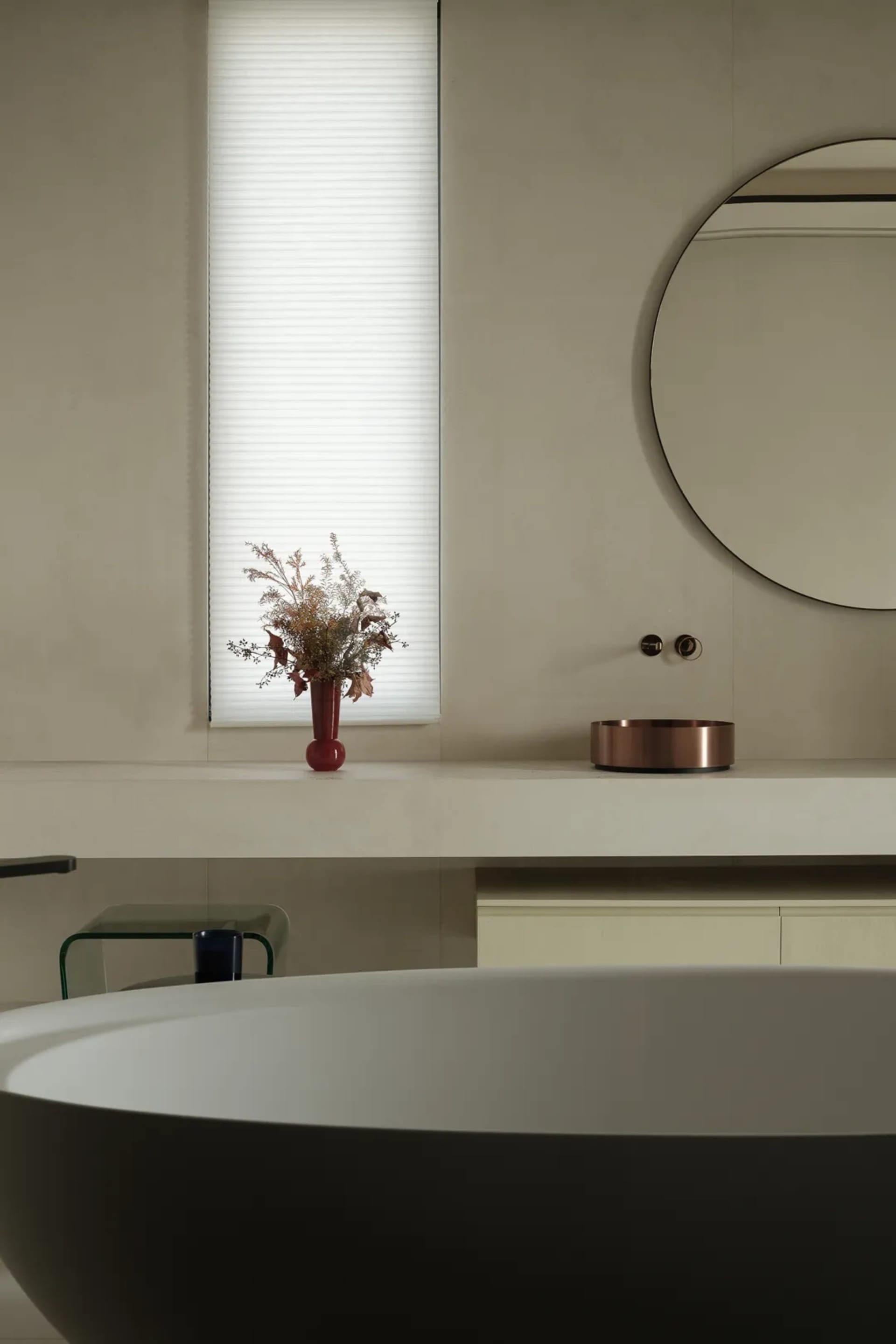 Modern minimalist bathroom interior with concrete walls, a round mirror, a vase with dried flowers, and a large window.