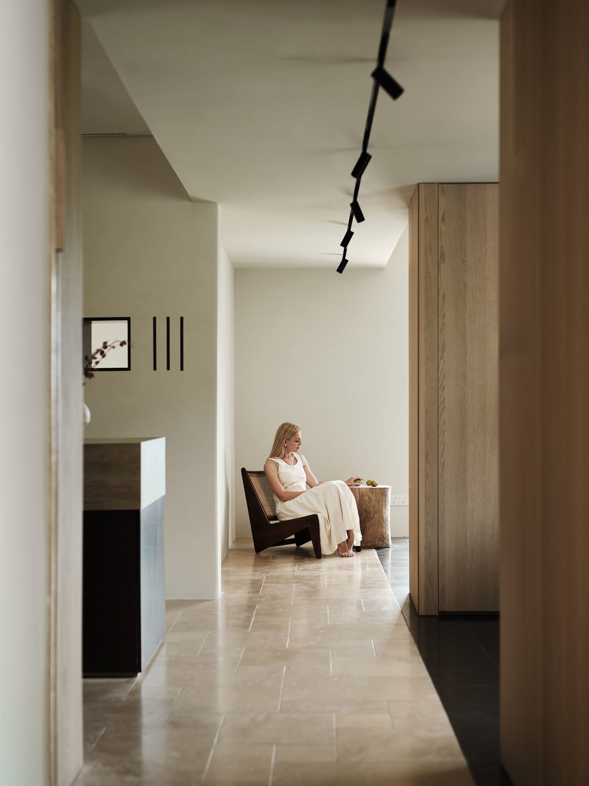 A woman in a white dress sits casually on a wooden chair in a modern residential space. Minimalist design with exposed concre