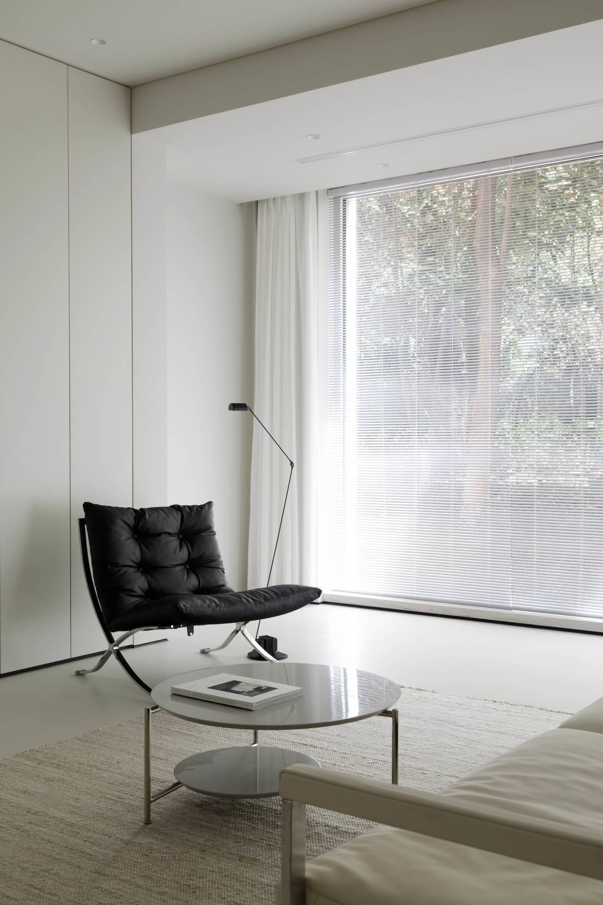 Modern minimalist living room with a black leather chair, white coffee table, and large window with blinds.