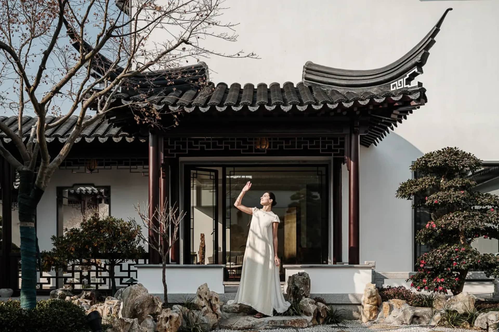 A woman in a white dress standing in front of a traditional Chinese pavilion with a rock garden and bonsai trees.