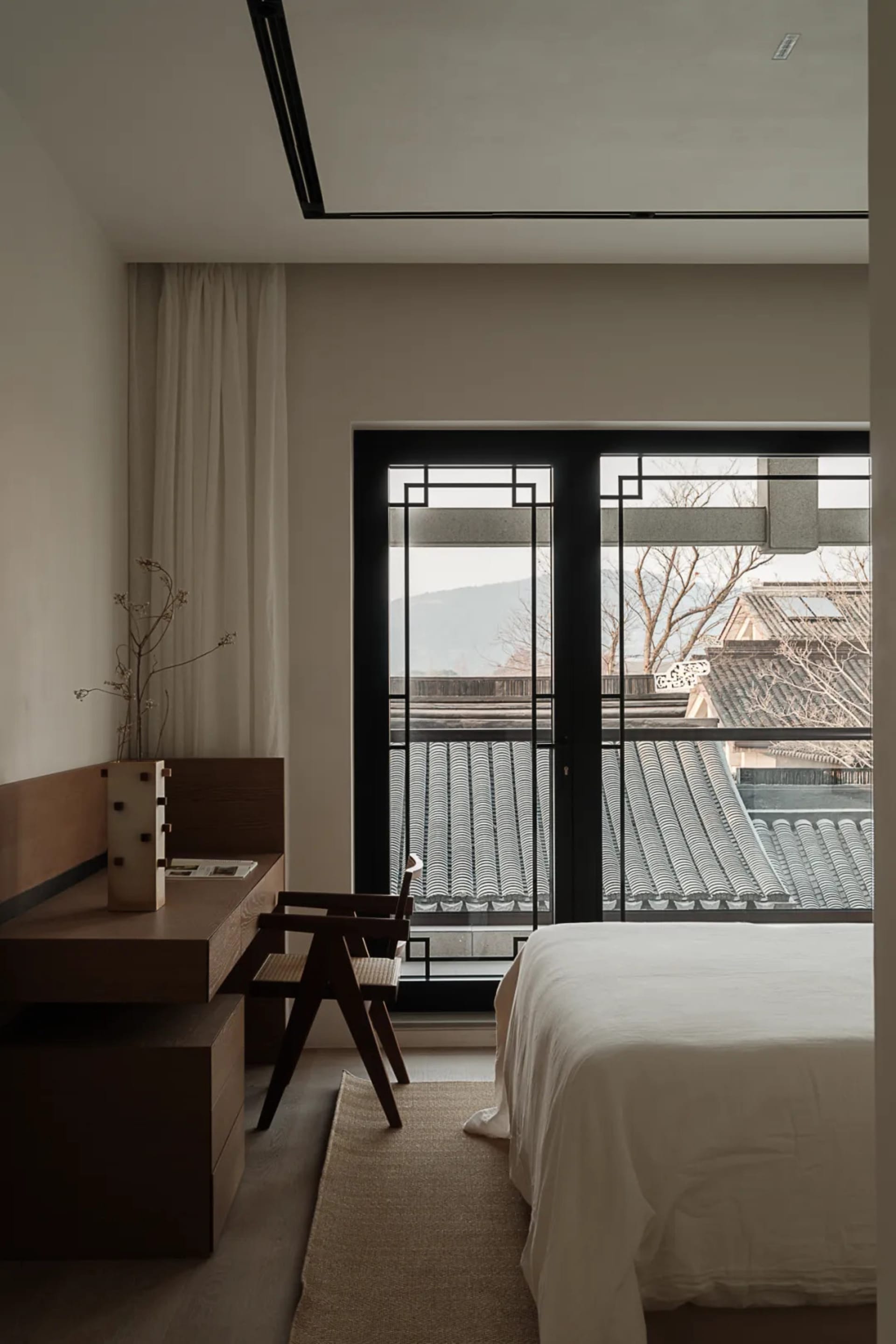 A modern minimalist bedroom with a white bed, wooden furniture, and a view of tiled rooftops.