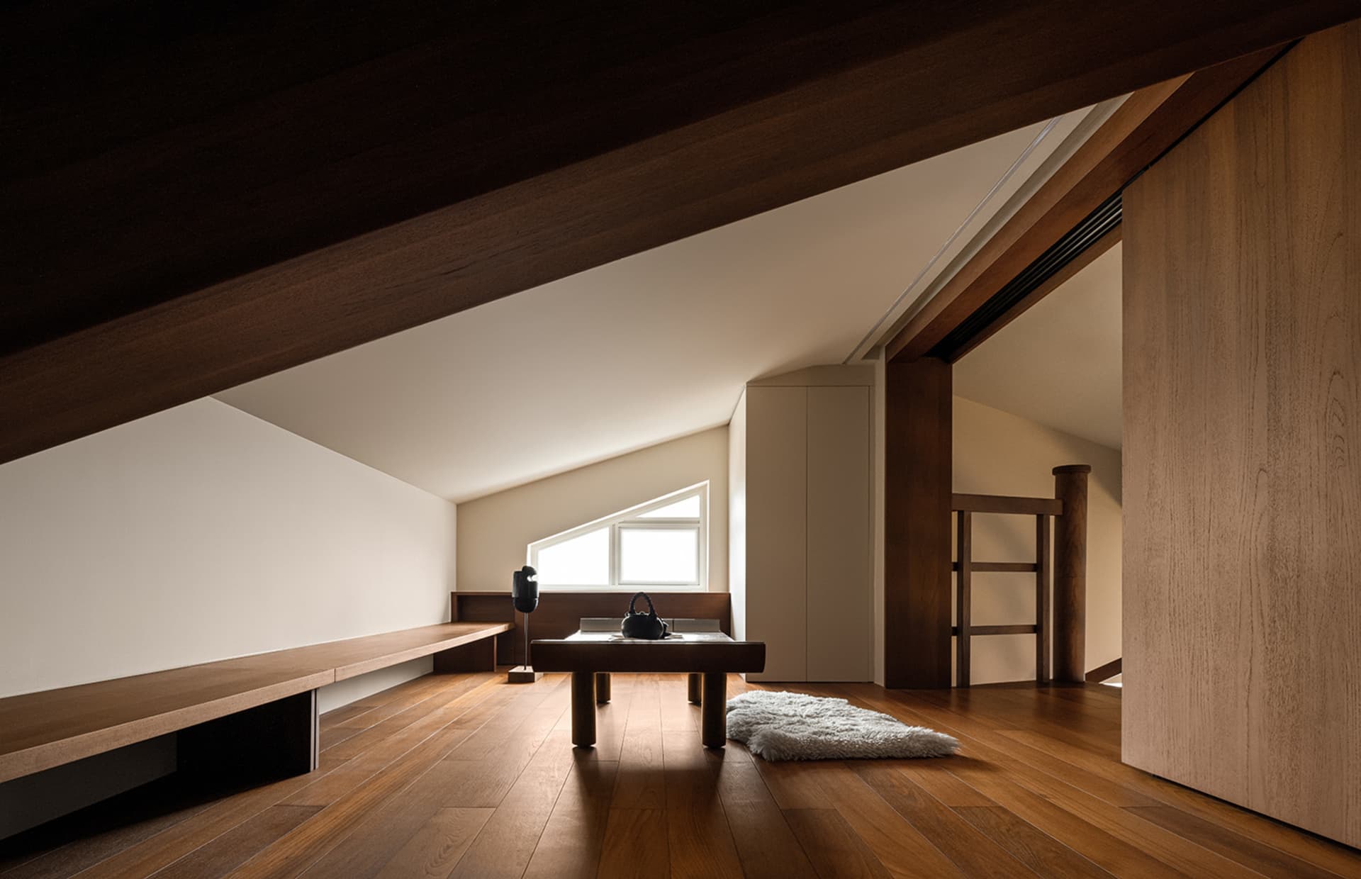 A minimalist living space with a wood-finished floor, featuring a bench, a table with a lamp, and a rug.