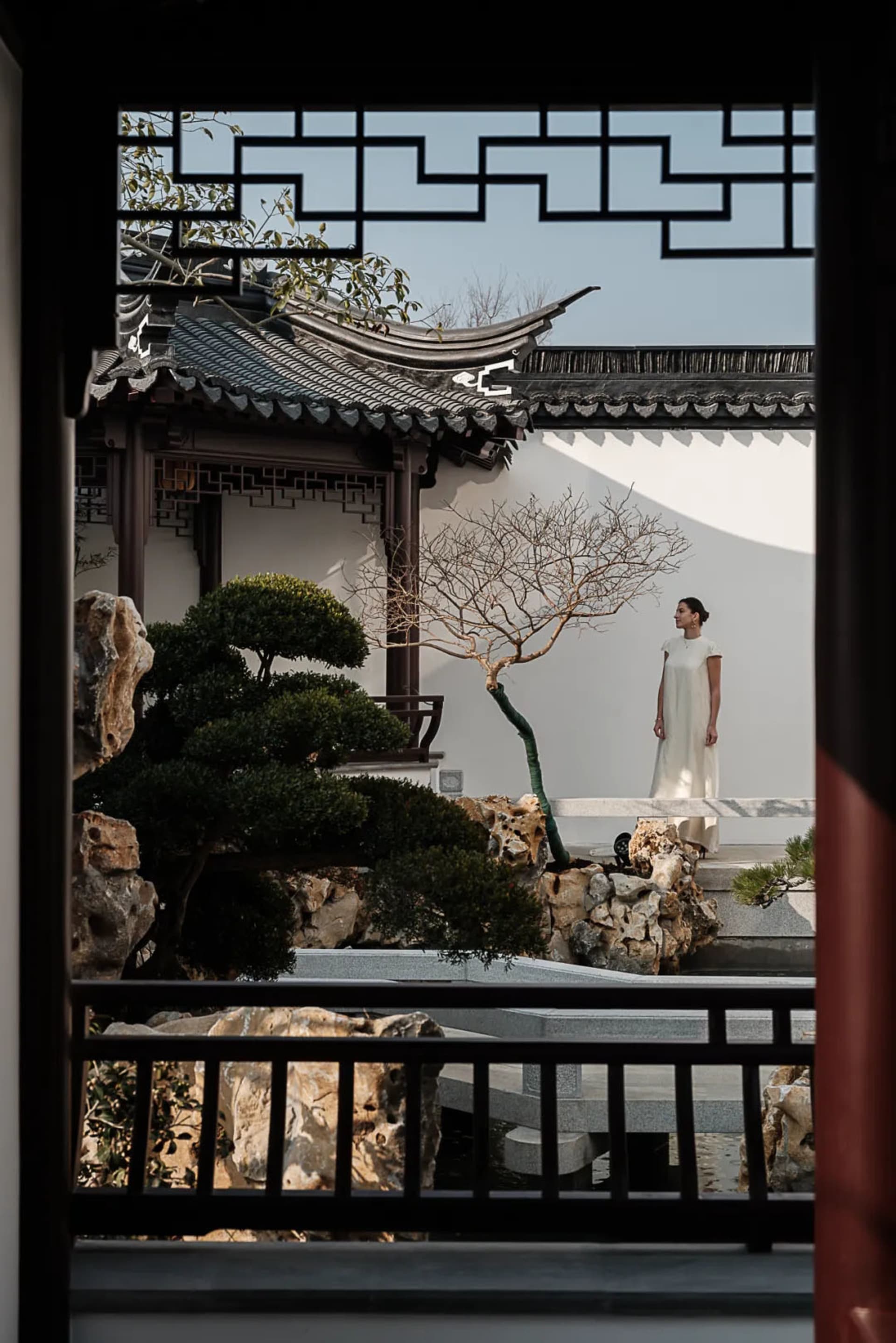 A person in a white dress standing in a traditional garden setting with stone, wooden structures, and glass elements.