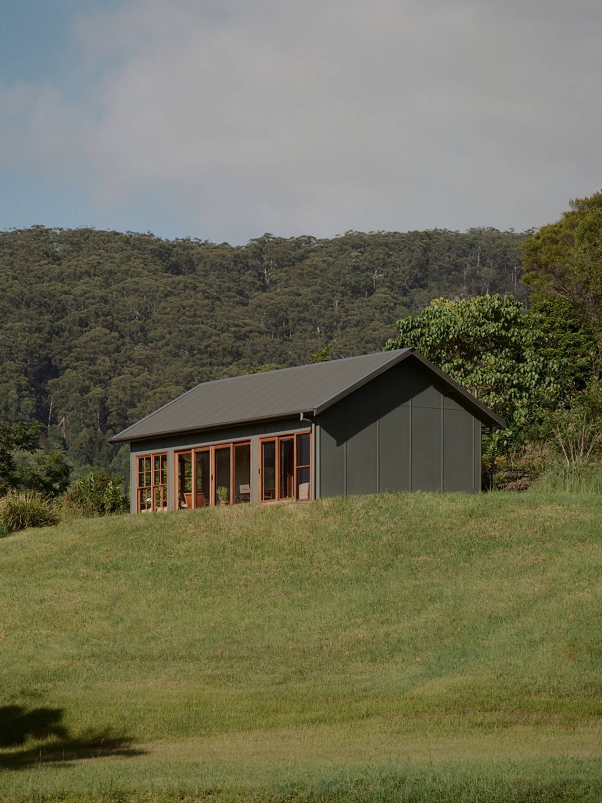A modern, minimalist house with a metal exterior, situated in a grassy field with a forested hill in the background.