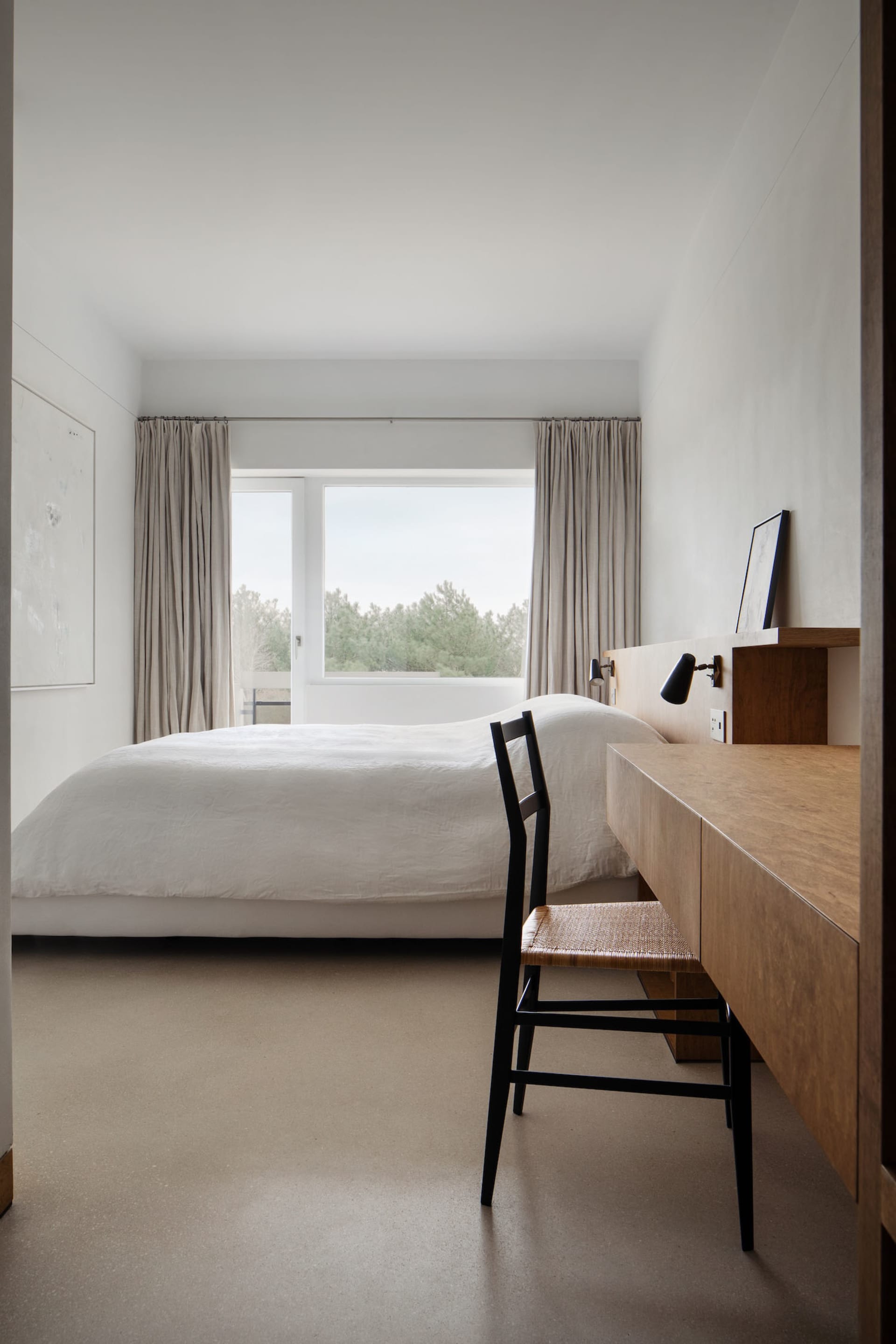 A minimalist bedroom featuring a wooden bed frame and matching desk, with light curtains and a view of trees through the wind