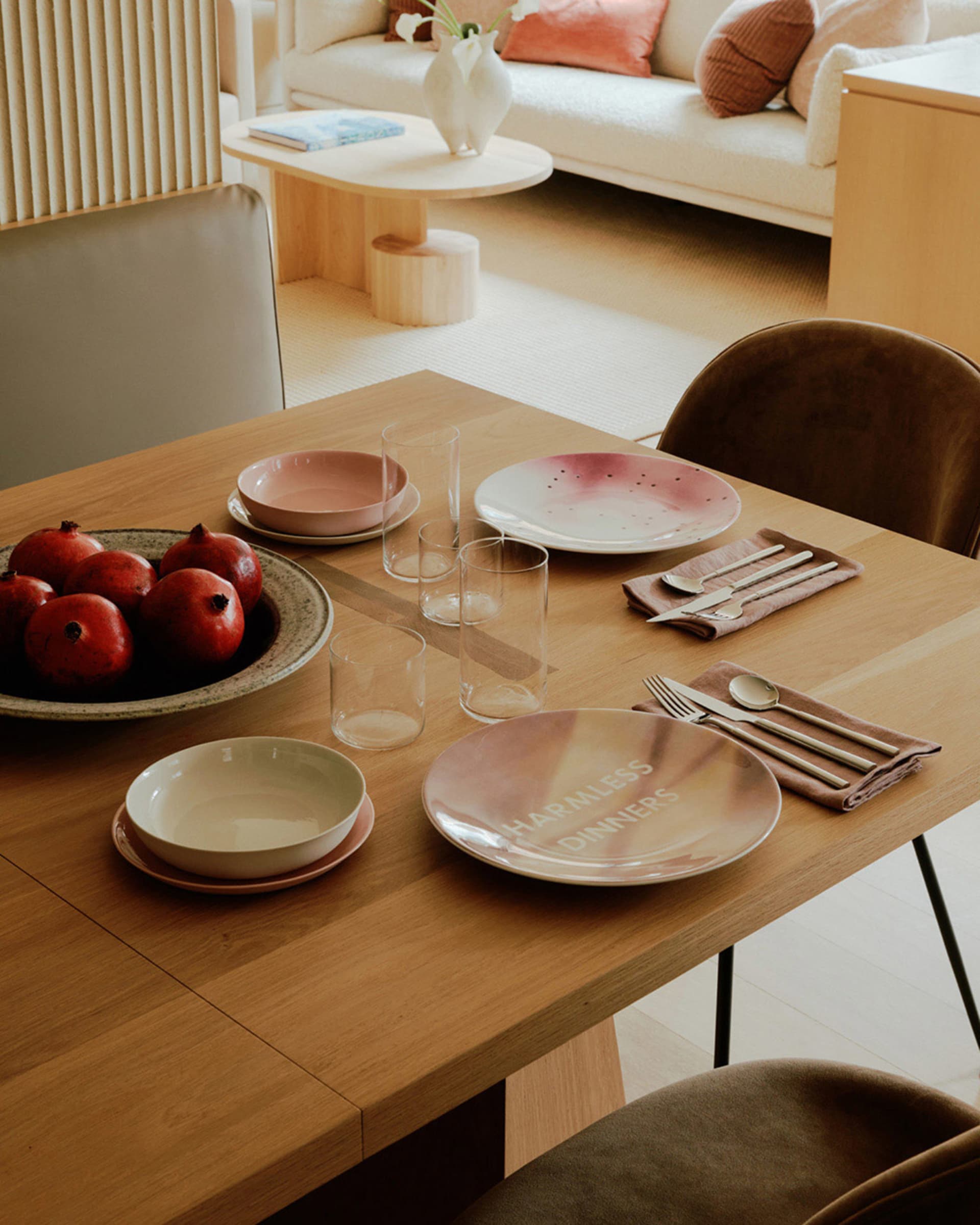 A modern minimalist dining room setup with wooden furniture, bowls, glasses, pink plates, cutlery, and pomegranates.