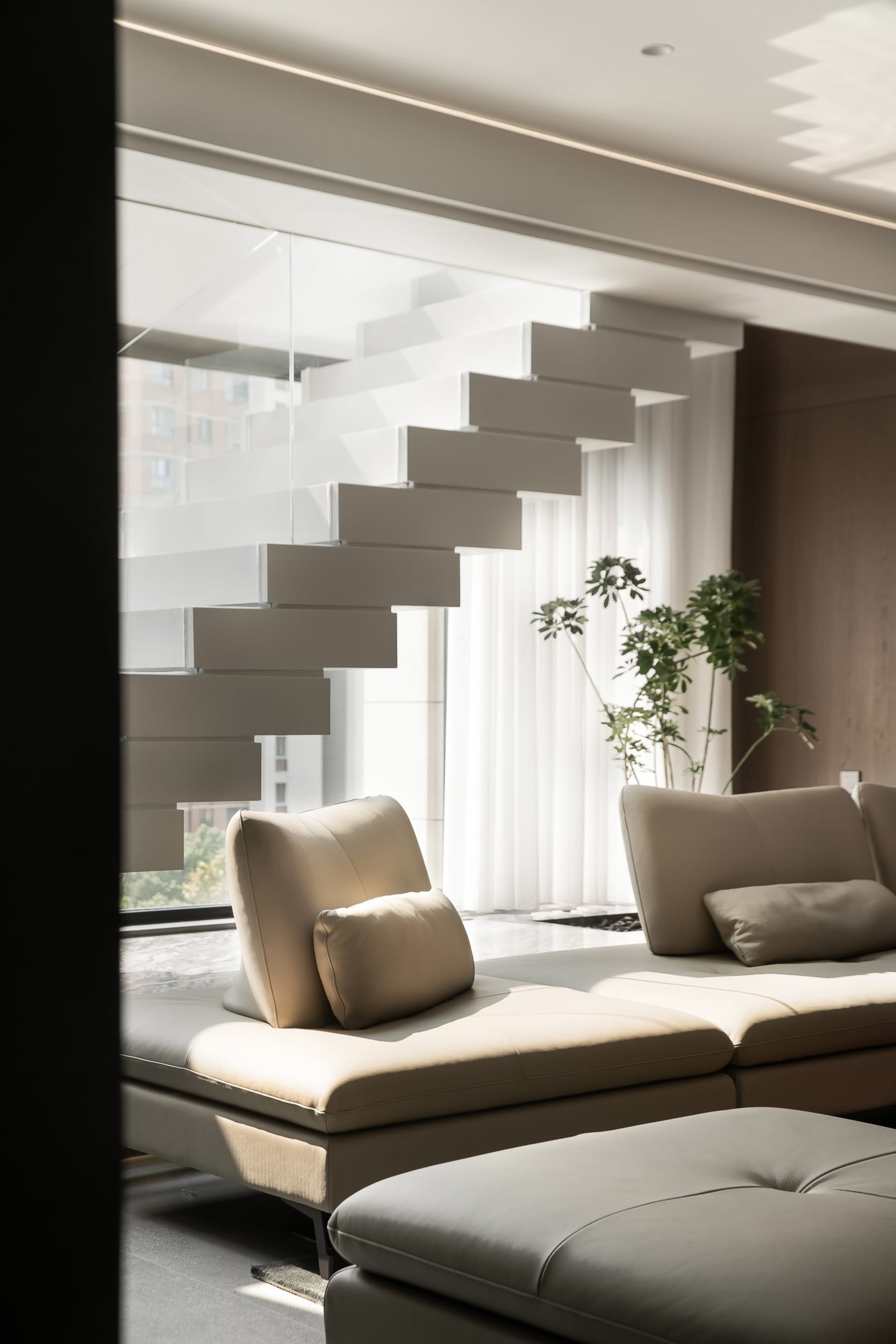 Modern living room with cream-colored leather furniture, glass staircases, and white curtains.