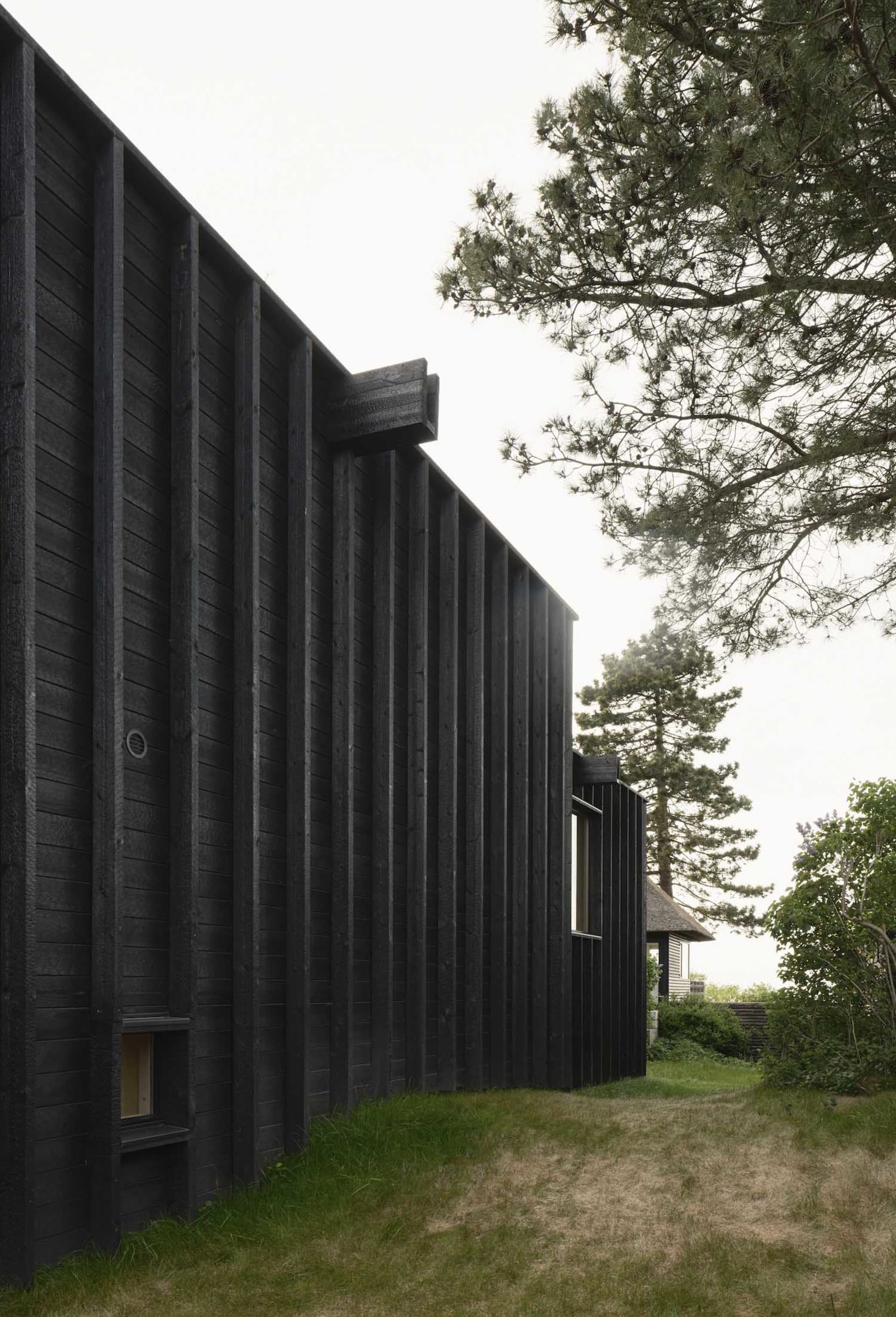 Black wooden exterior of a modern house with a dirt path and trees in the background.