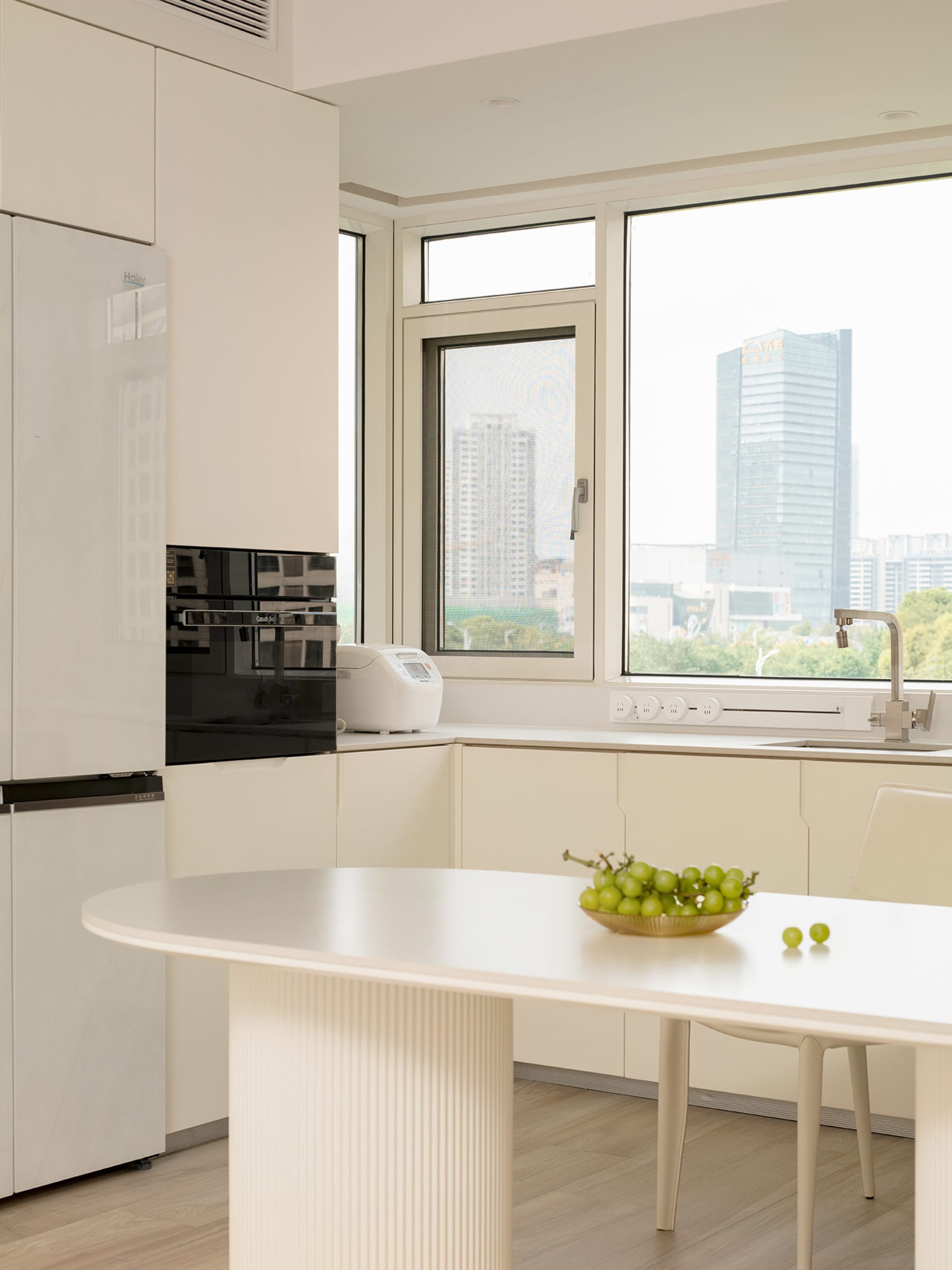 A modern minimalist kitchen featuring white laminate cabinets, a sleek countertop, and a view through a large window.