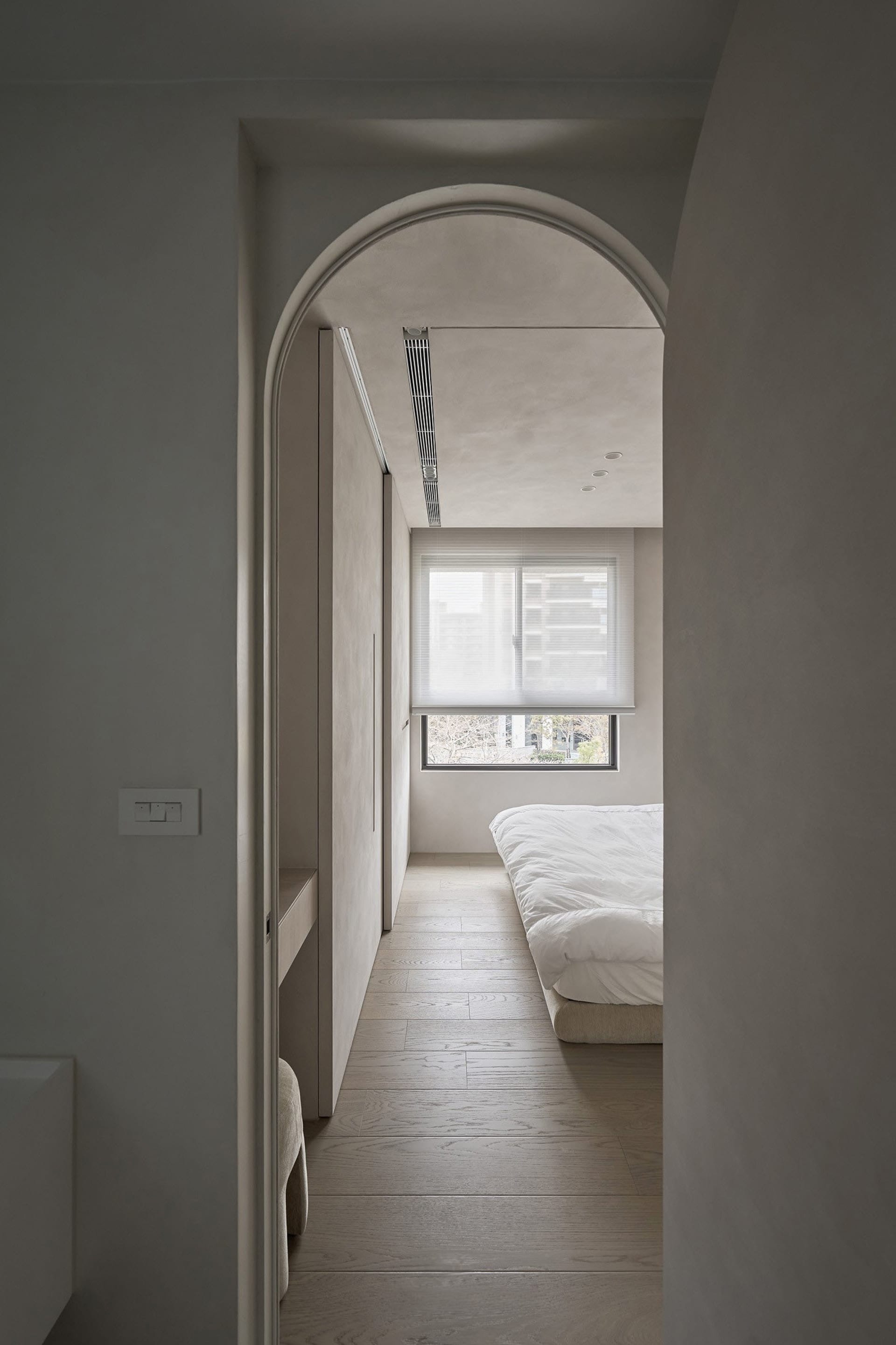 A minimalist bedroom featuring light-colored wood flooring, white bedding, and a large window with blinds.