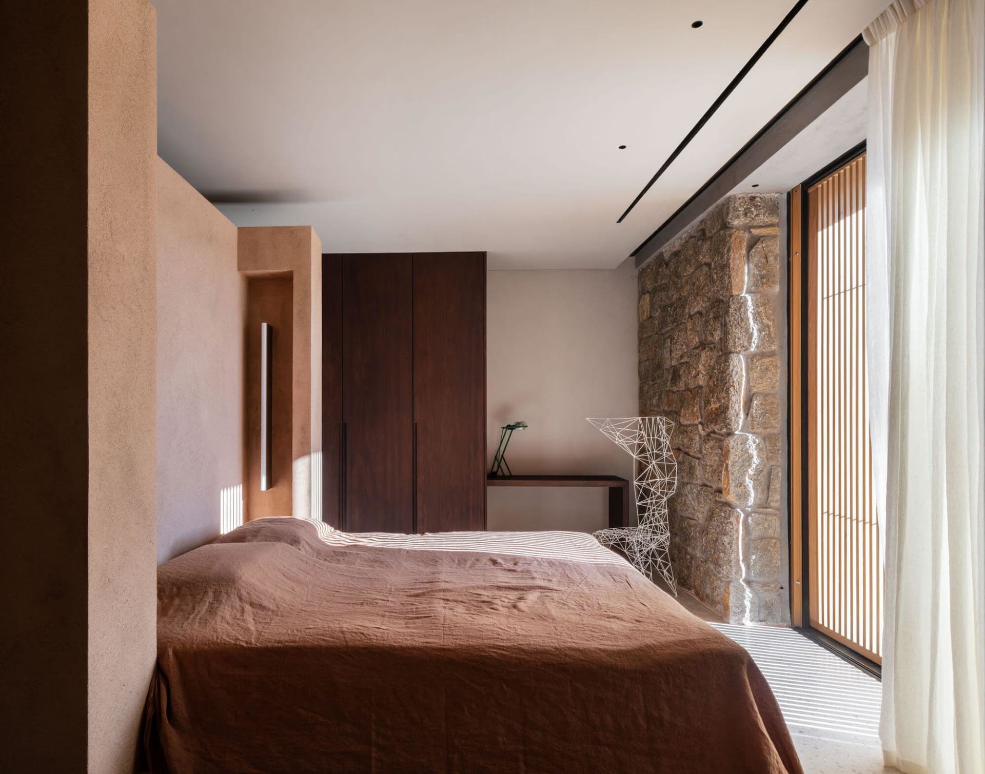 A minimalist bedroom featuring wooden furniture and a stone wall, with natural light streaming through the window.