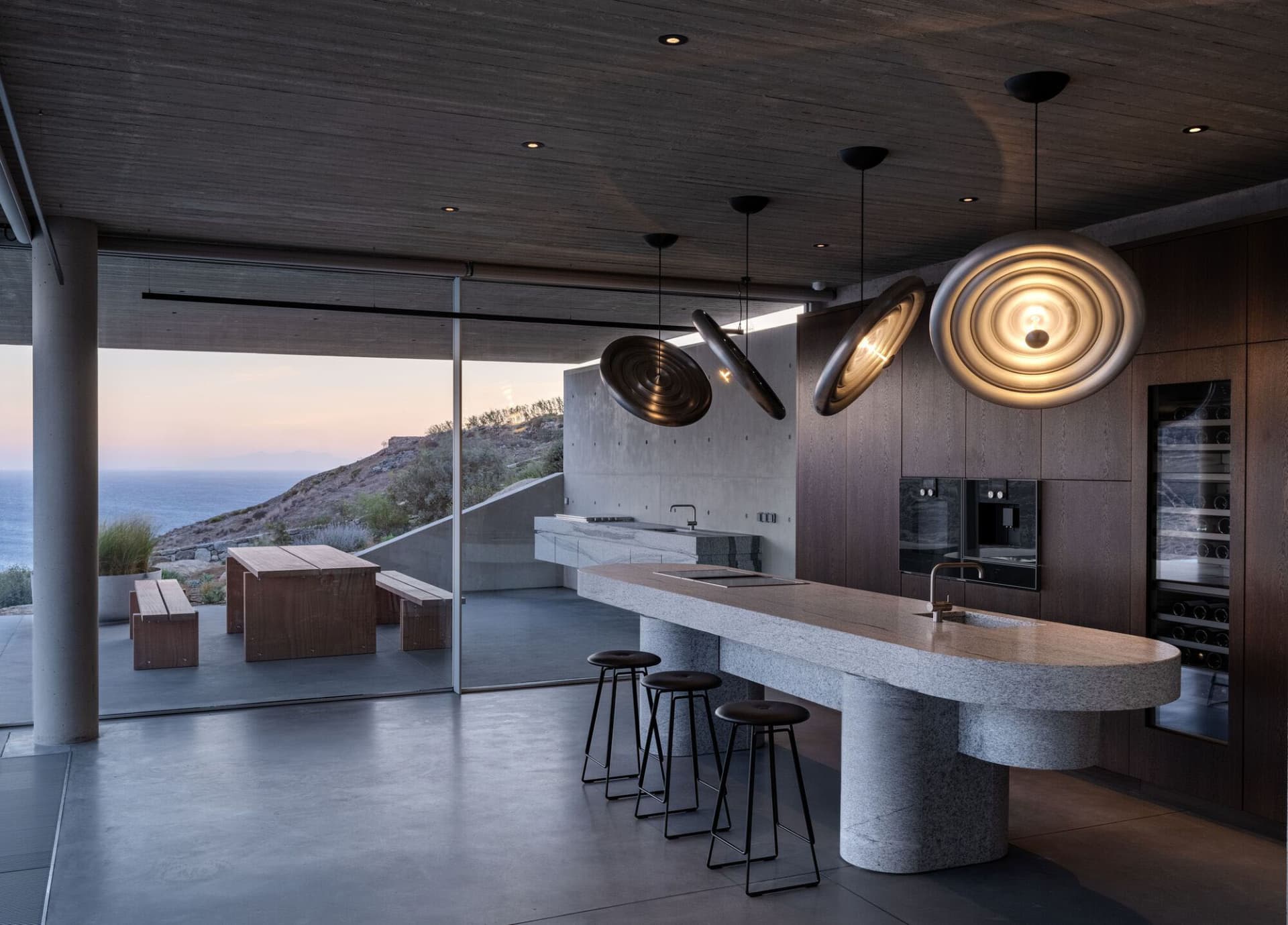 A modern industrial-style kitchen featuring granite countertops and a concrete ceiling, with a view of the ocean and mountain