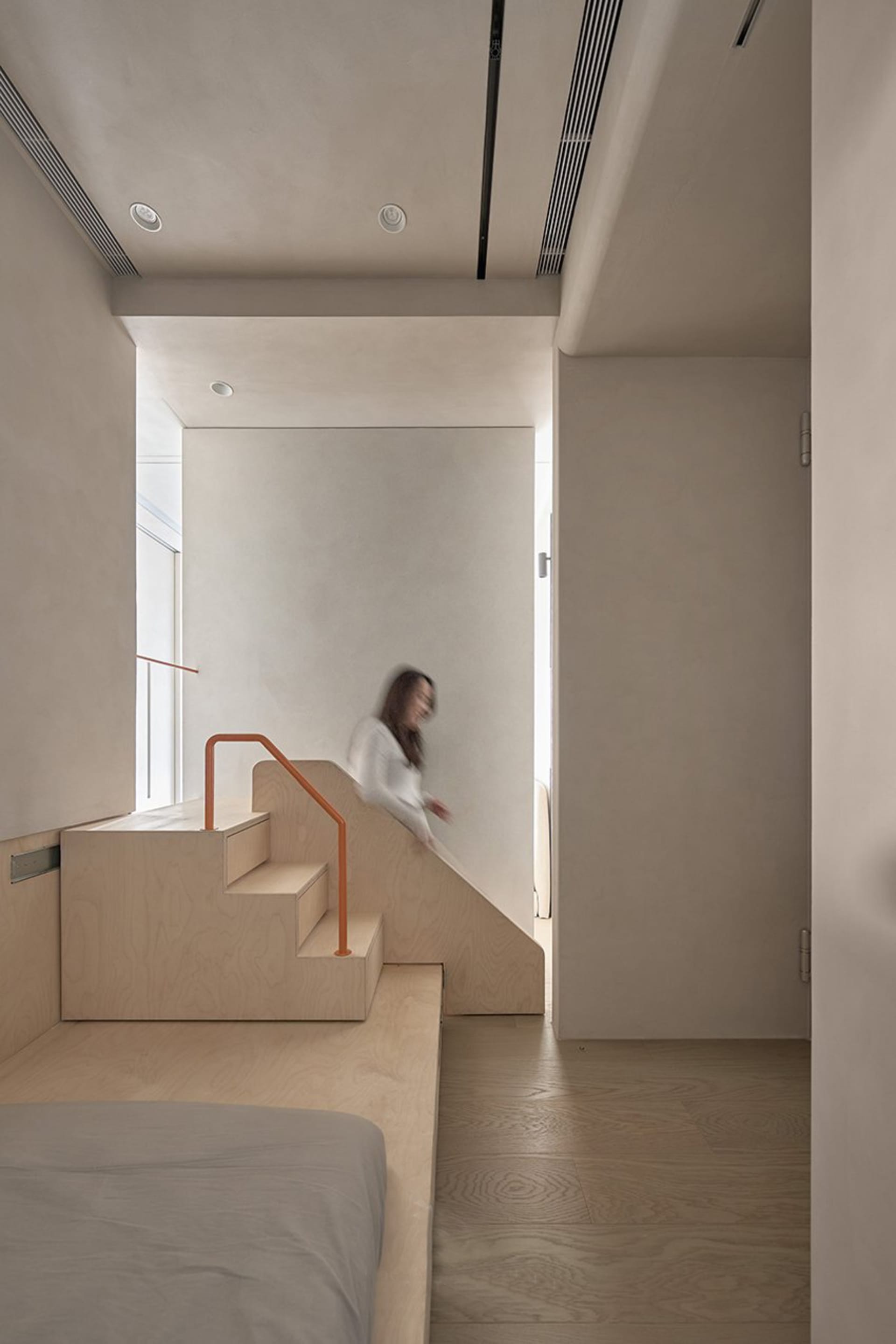 A minimalist interior space featuring wood and concrete elements, with a blurred figure walking down a staircase.