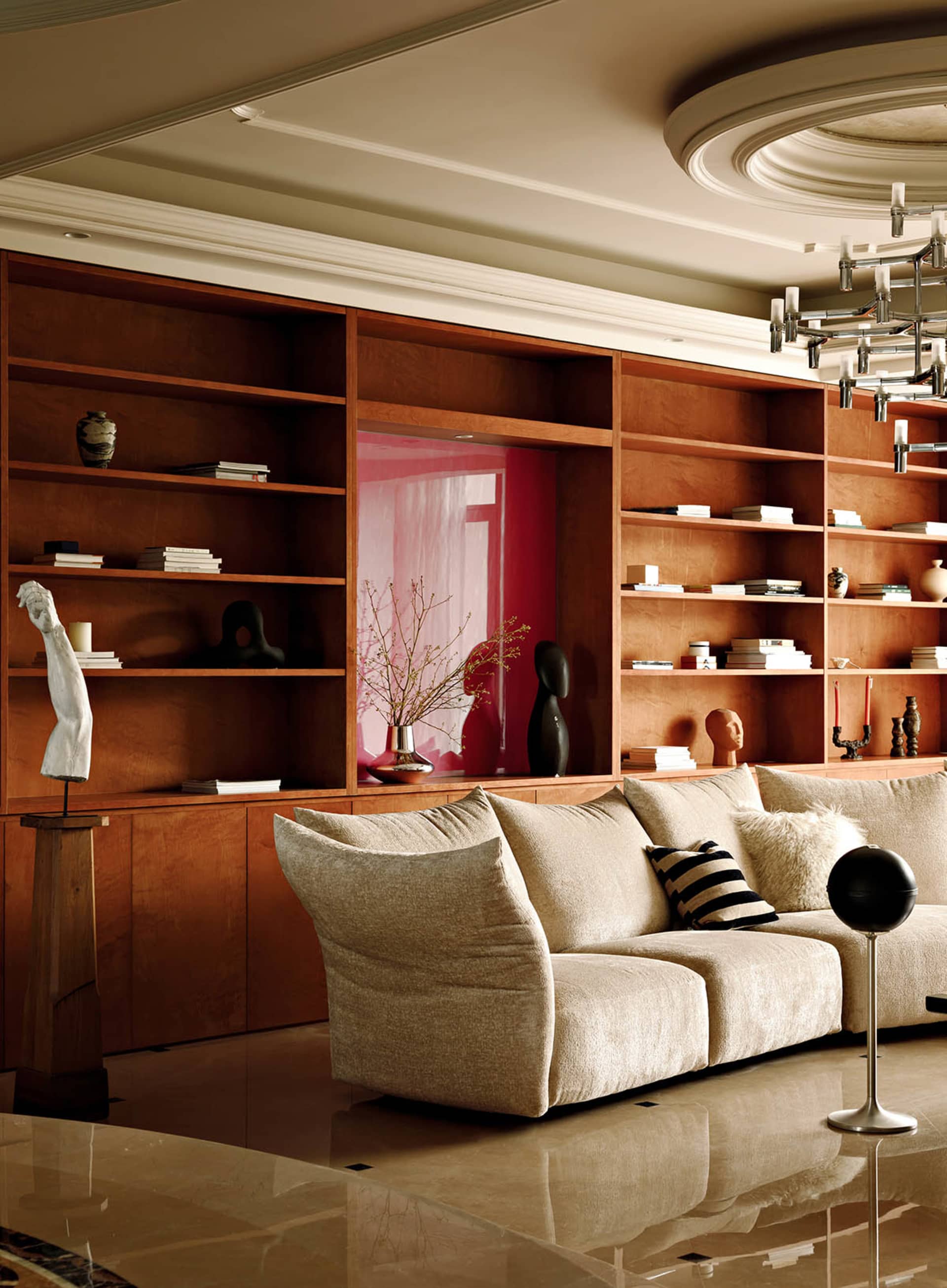 A modern living room with a large beige sofa, wooden shelves filled with books and decorative items, and a marble floor.