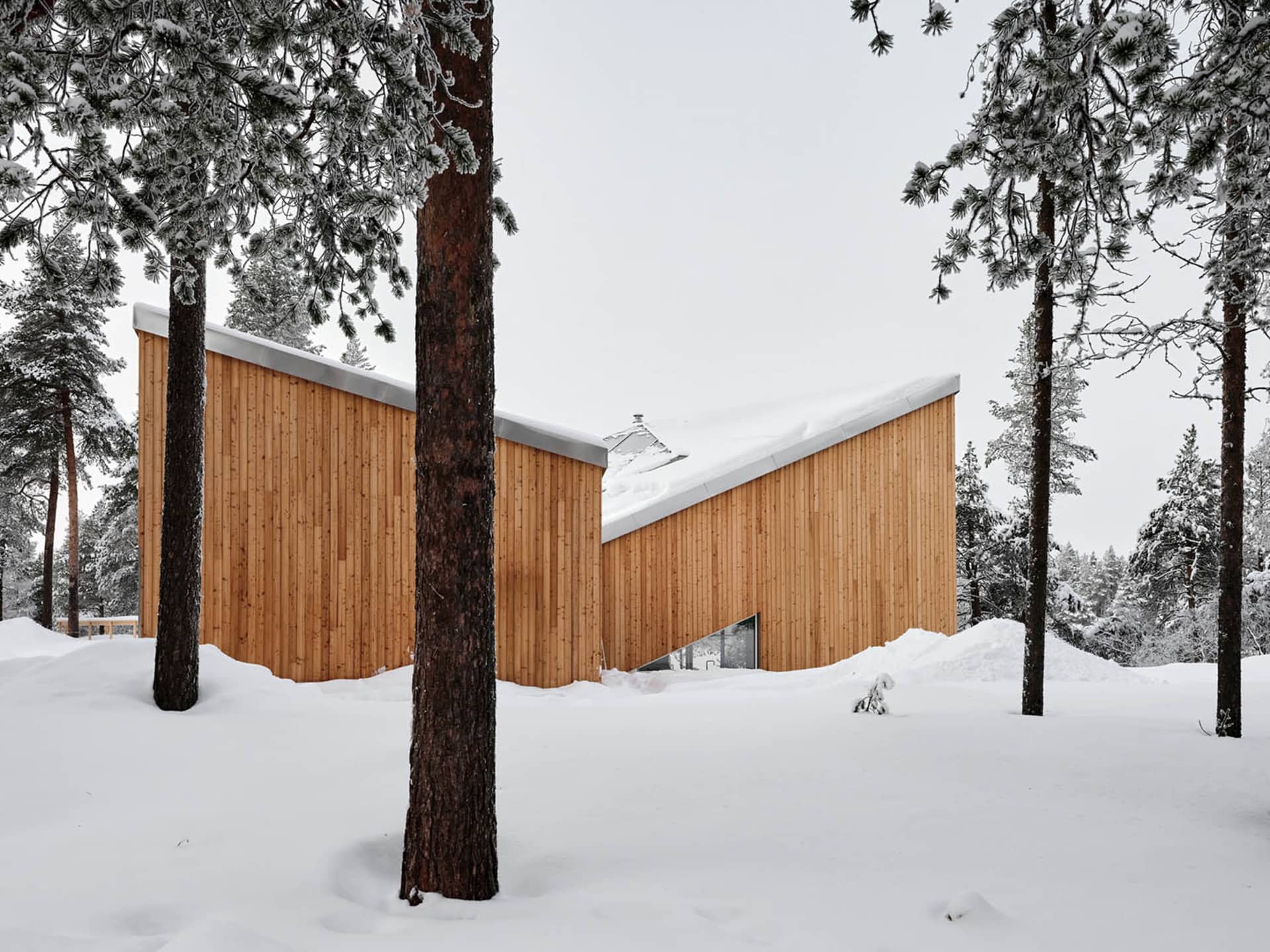 A modern wooden house sits in a winter landscape surrounded by snow-covered trees.