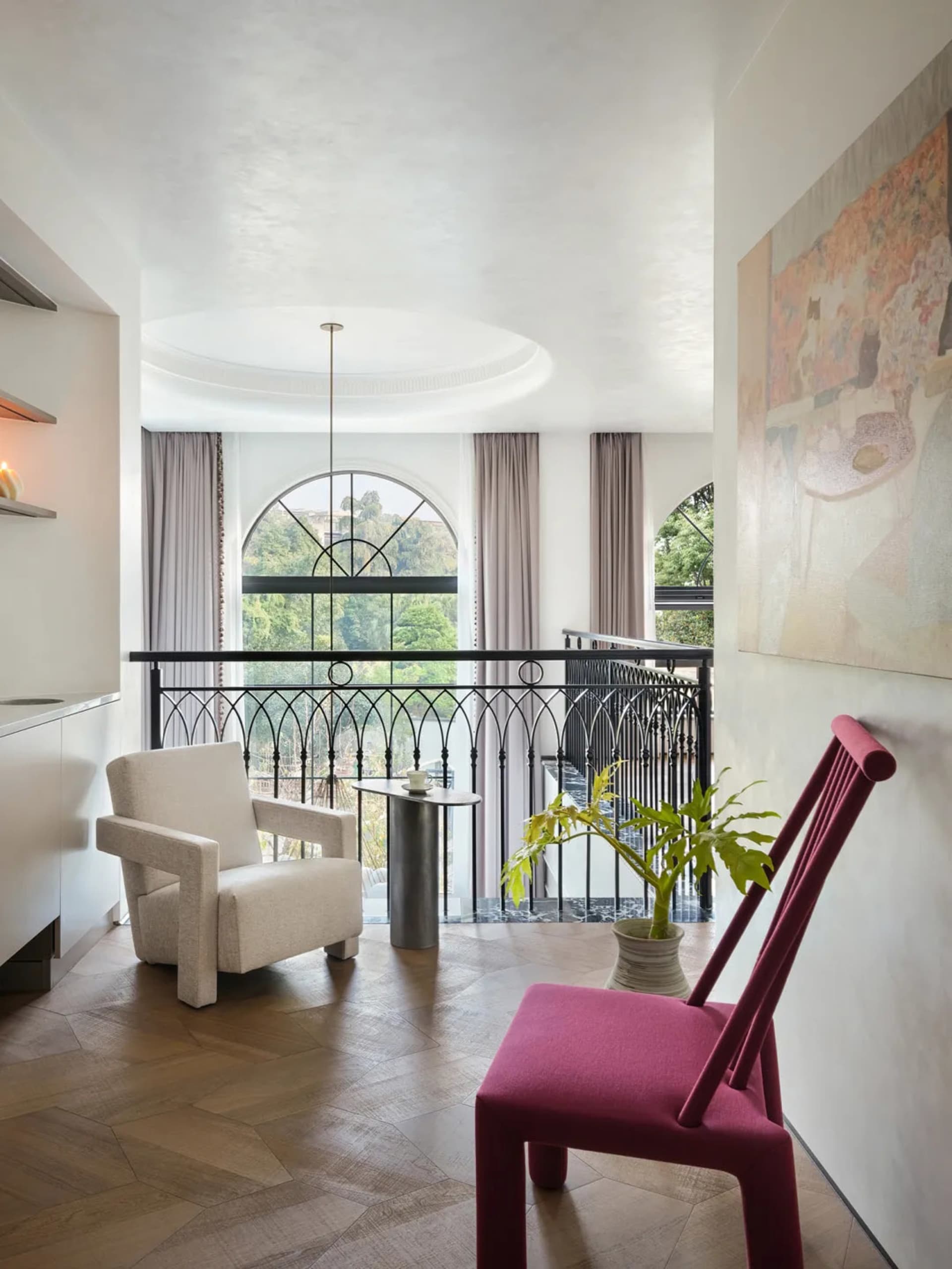 A modern living room with wooden furniture, a white armchair, a pink chair, and a large arched window.