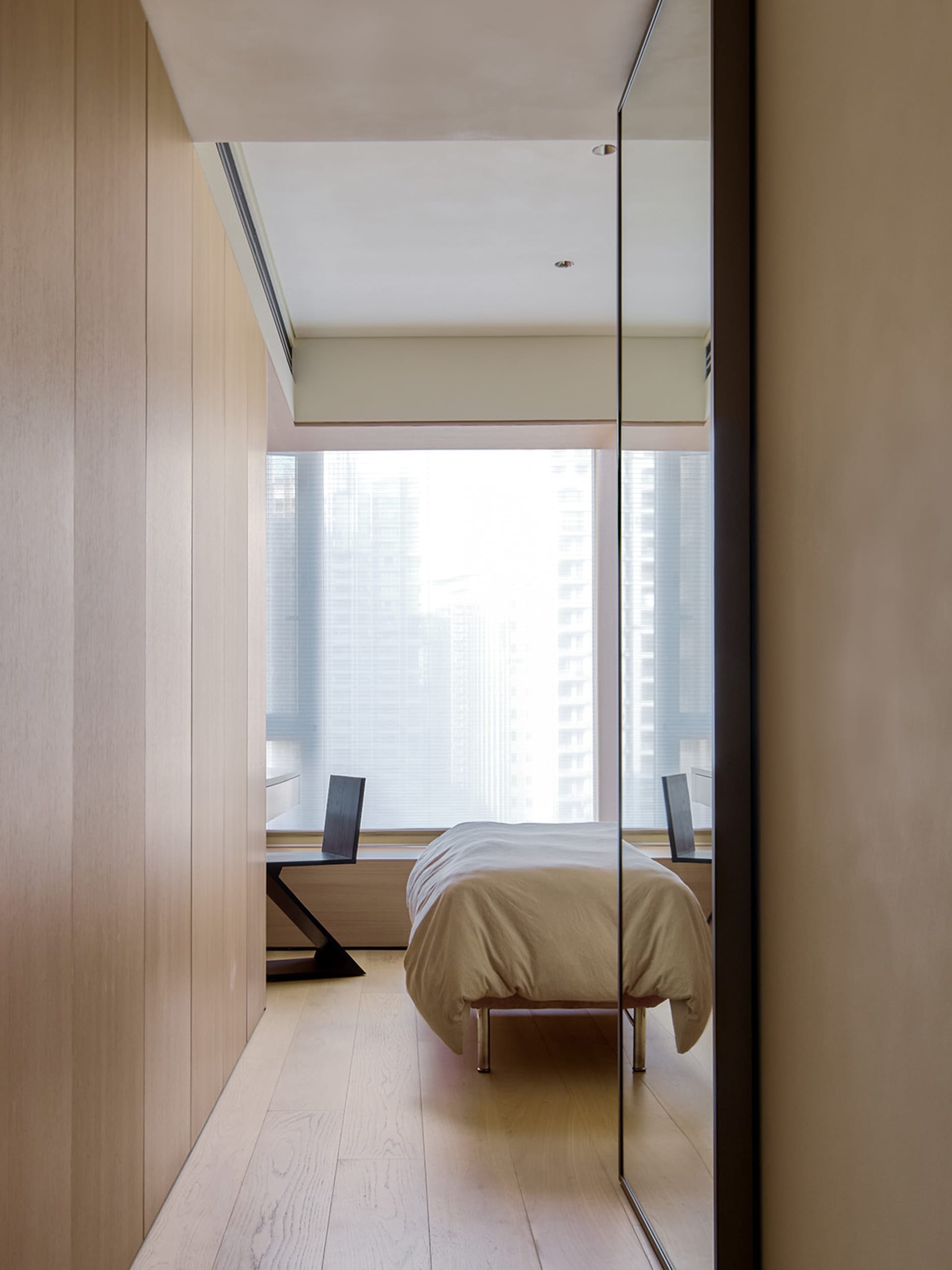 Modern minimalist bedroom with light wood floors, glass window overlooking cityscape, and metal furniture.