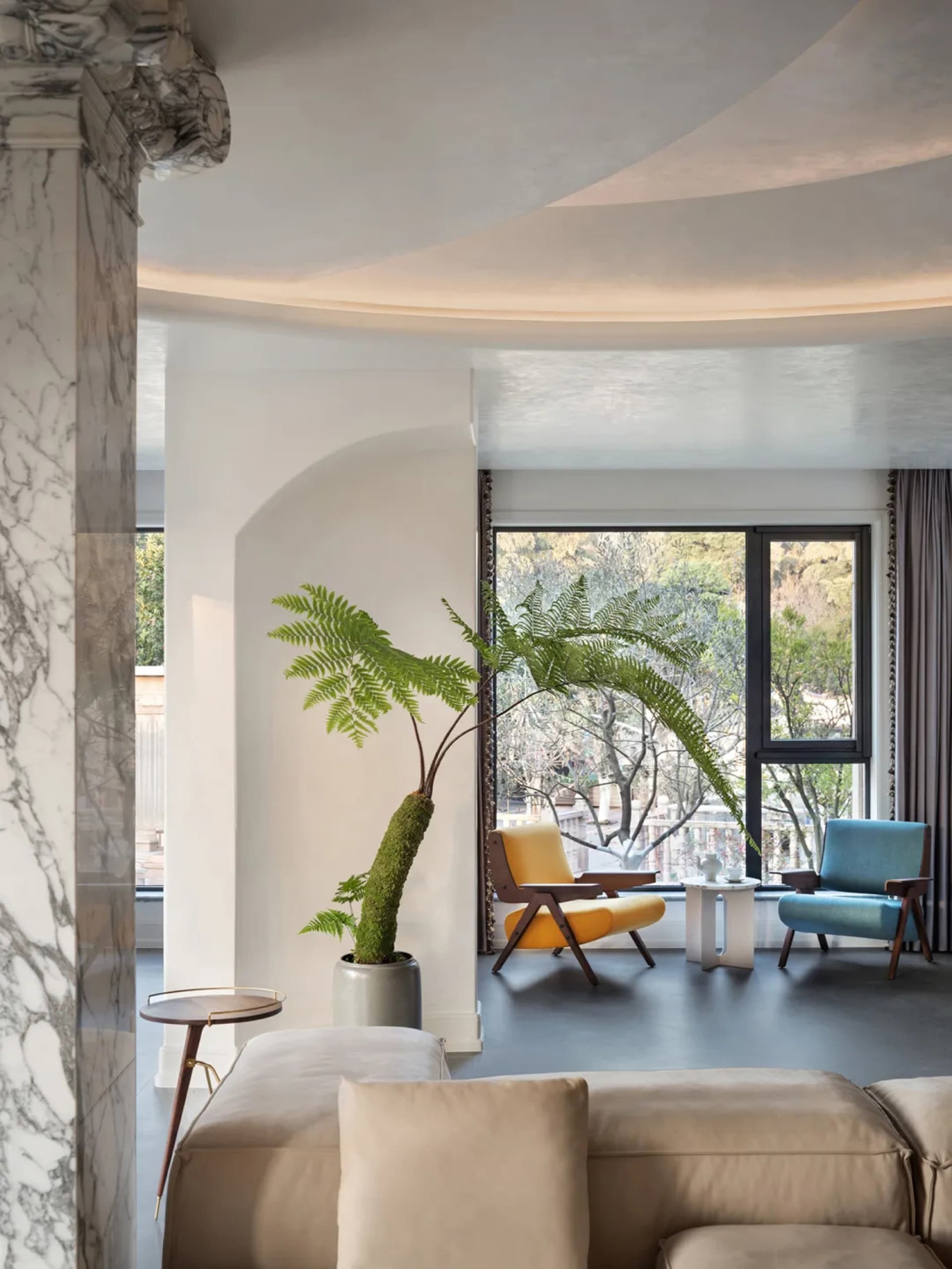 A modern minimalist living room with concrete walls, a beige sofa, a yellow chair, a blue chair, and a potted fern plant.