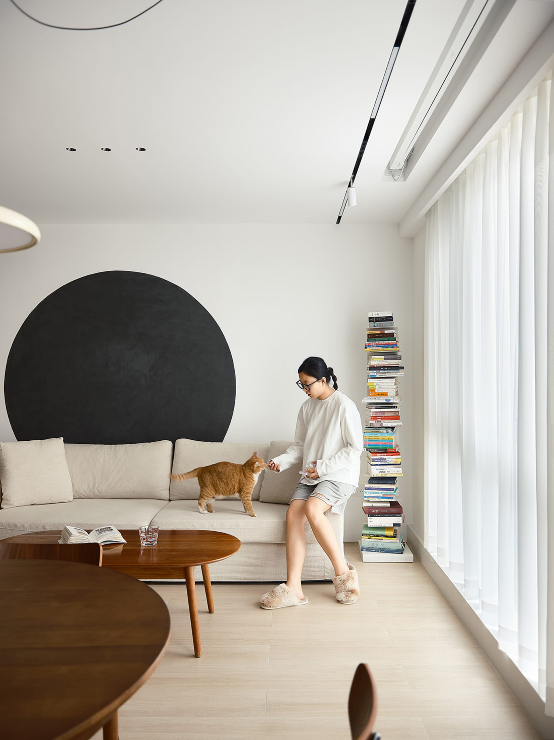 A minimalist living room with a beige sectional sofa, wooden tables, and a black circular accent on the wall.