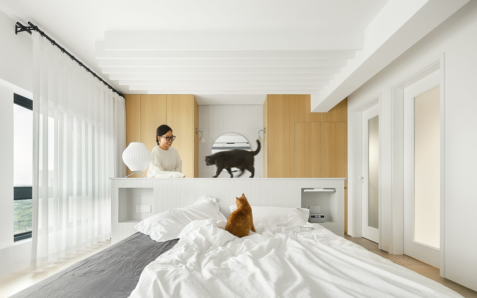 A modern minimalistic bedroom featuring white bedding and curtains, and a black cat on a wooden counter.