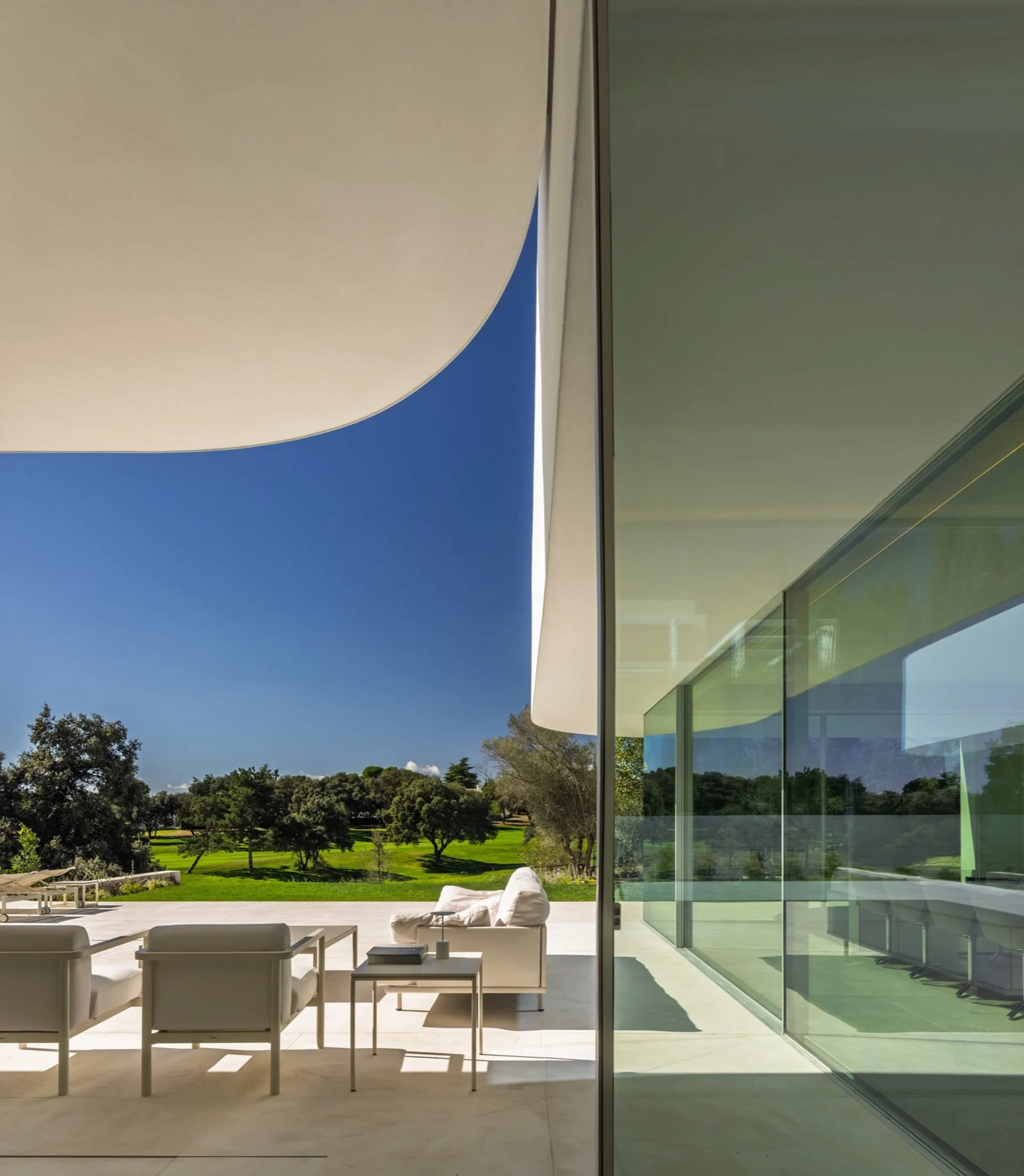 Modern outdoor lounge area featuring glass walls and a concrete floor, overlooking greenery and clear blue sky.