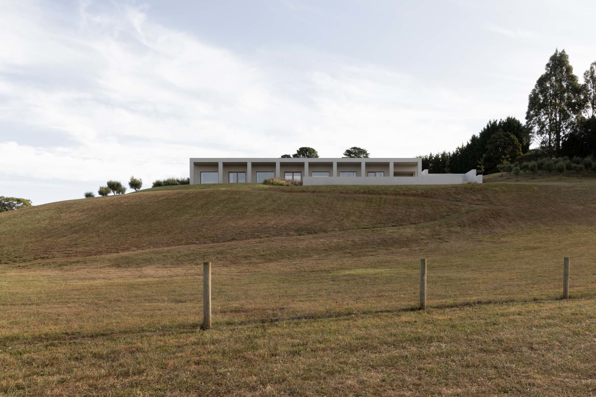 A sleek, modern house with concrete and glass materials sits atop a grassy hillside.