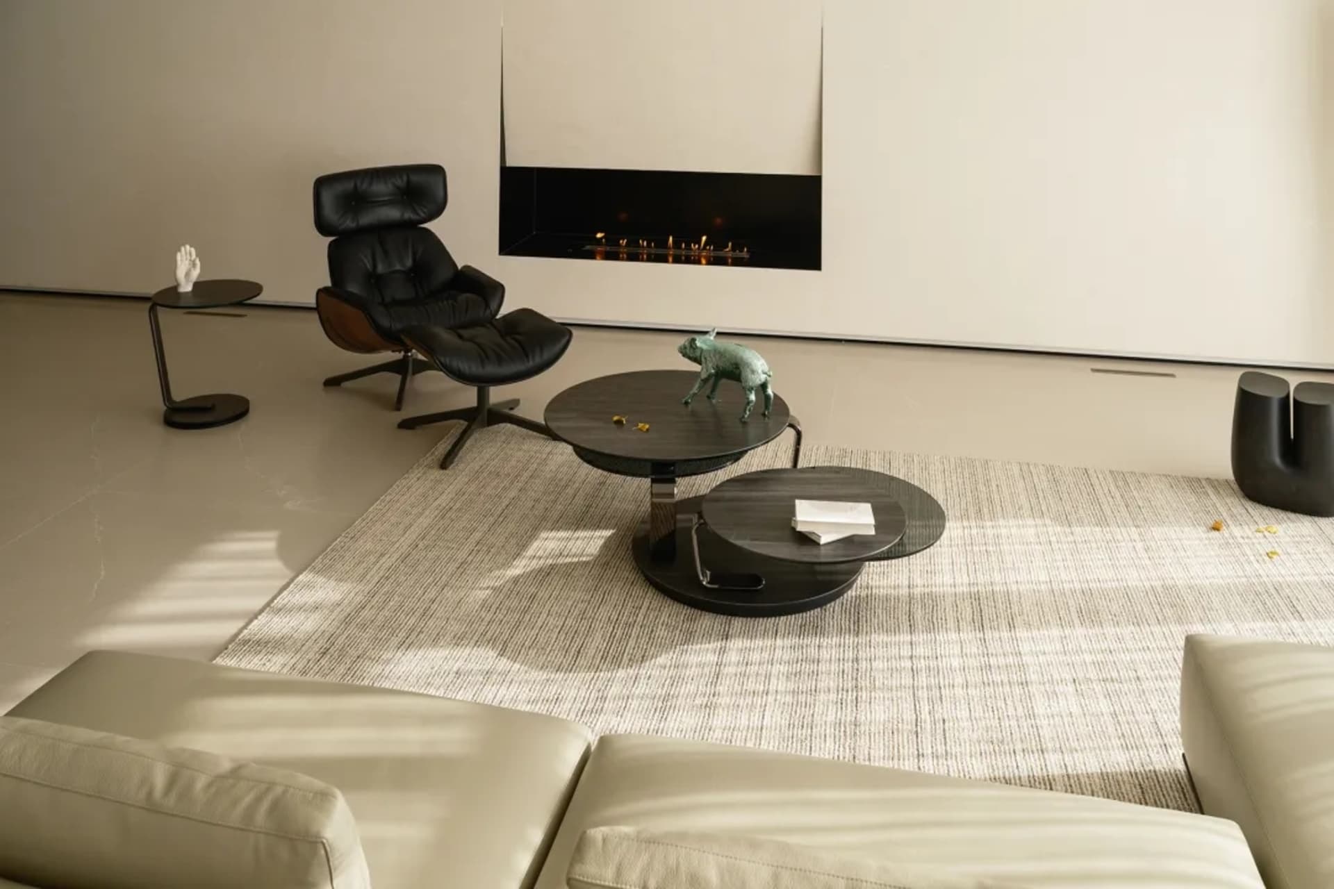 A minimalist living room featuring a modern black lounge chair, round black tables with books and figurines, a beige sofa, an