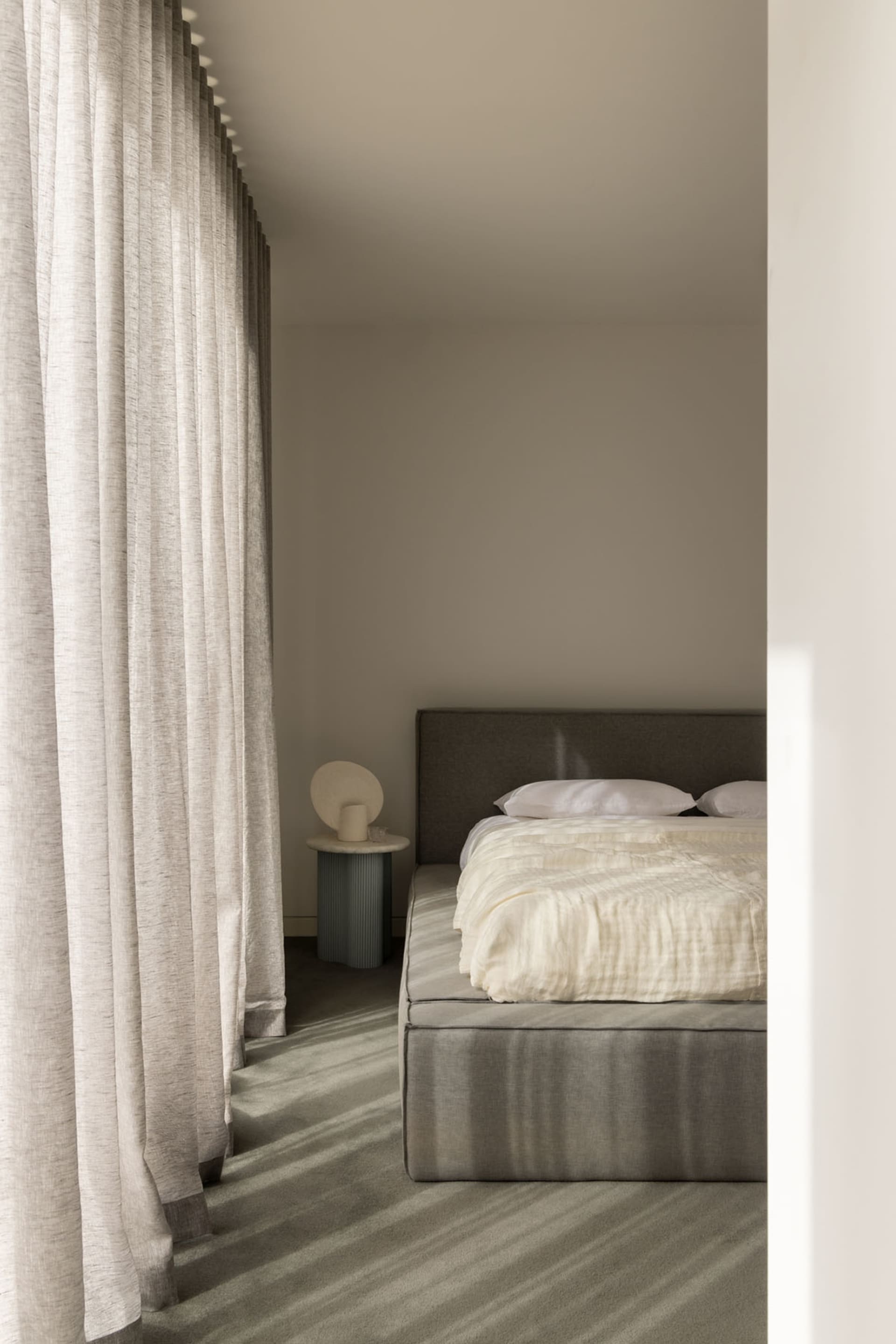 A modern bedroom featuring a grey upholstered bed with white bedding, surrounded by sheer curtains and a textured grey carpet